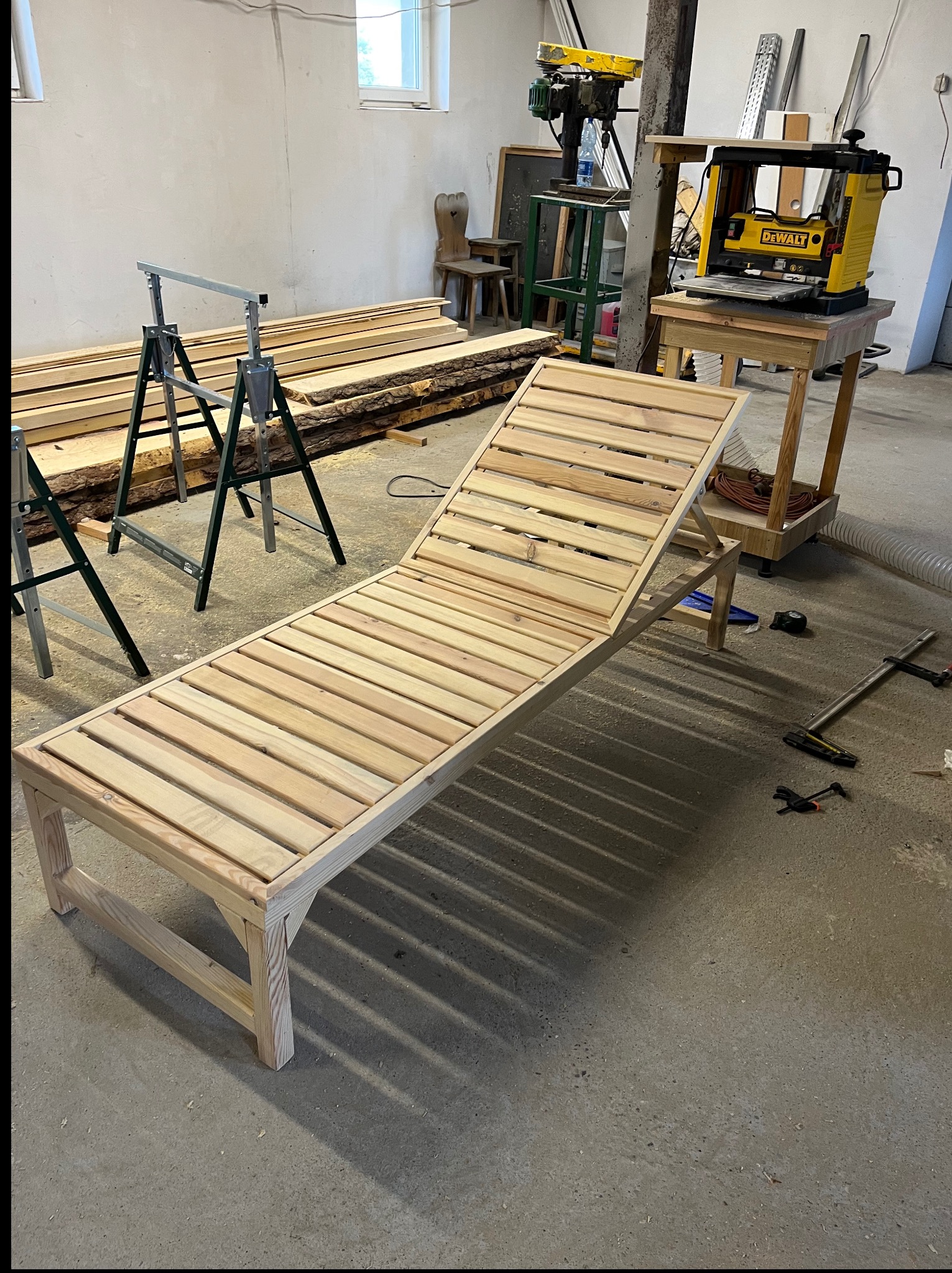 Drewniany leżak ogrodowy w warsztacie, przed obróbką. W tle strugarka, piła i deski. Widoczne narzędzia stolarskie. Surowy, naturalny wygląd drewna.