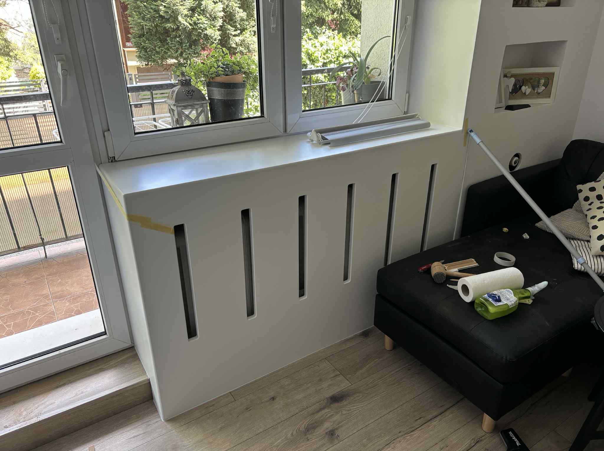 Biała obudowa grzejnika z pionowymi szczelinami, zamontowana pod oknem balkonowym. Na pufie narzędzia i akcesoria montażowe. Podłoga z paneli drewnopodobnych.