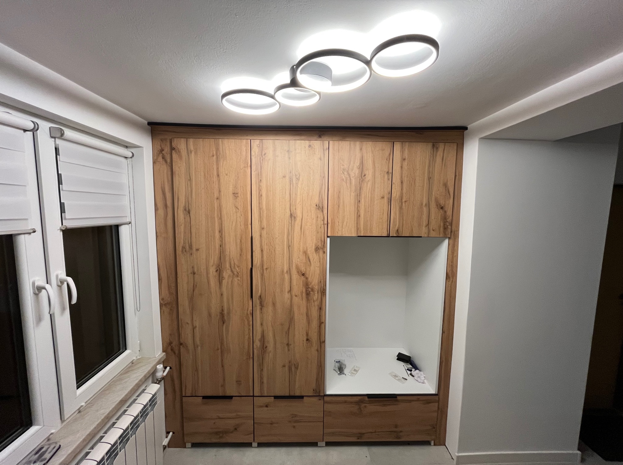 Drewniana szafa z otwartą wnęką, nowoczesne oświetlenie sufitowe. Widoczne detale wykończenia i akcesoria wewnątrz szafy. Minimalistyczny design.