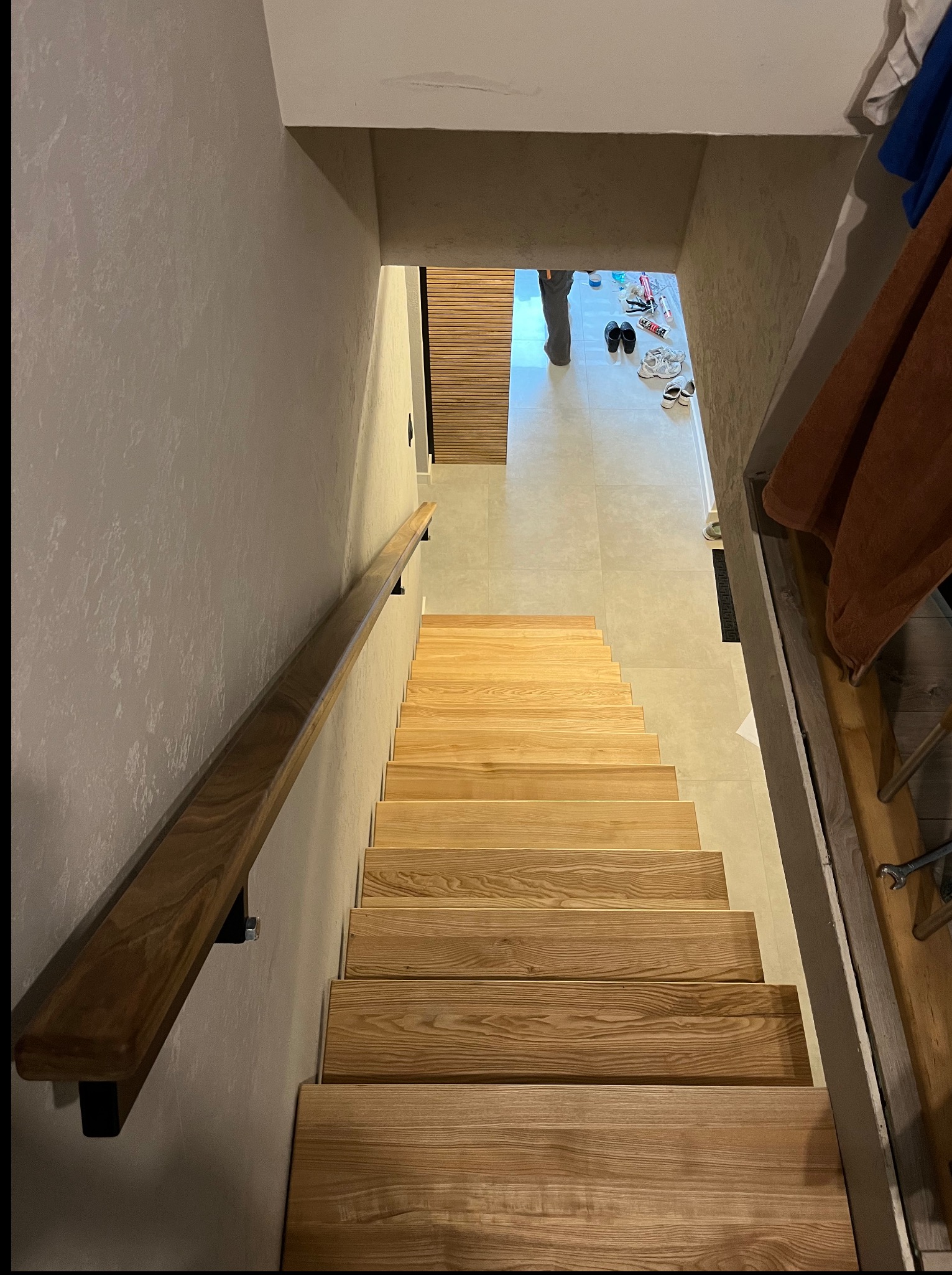Drewniane schody z poręczą, widok z góry. Naturalne wykończenie drewna, jasne stopnie. W tle fragment korytarza z butami i narzędzia. Ujęcie skierowane w dół.