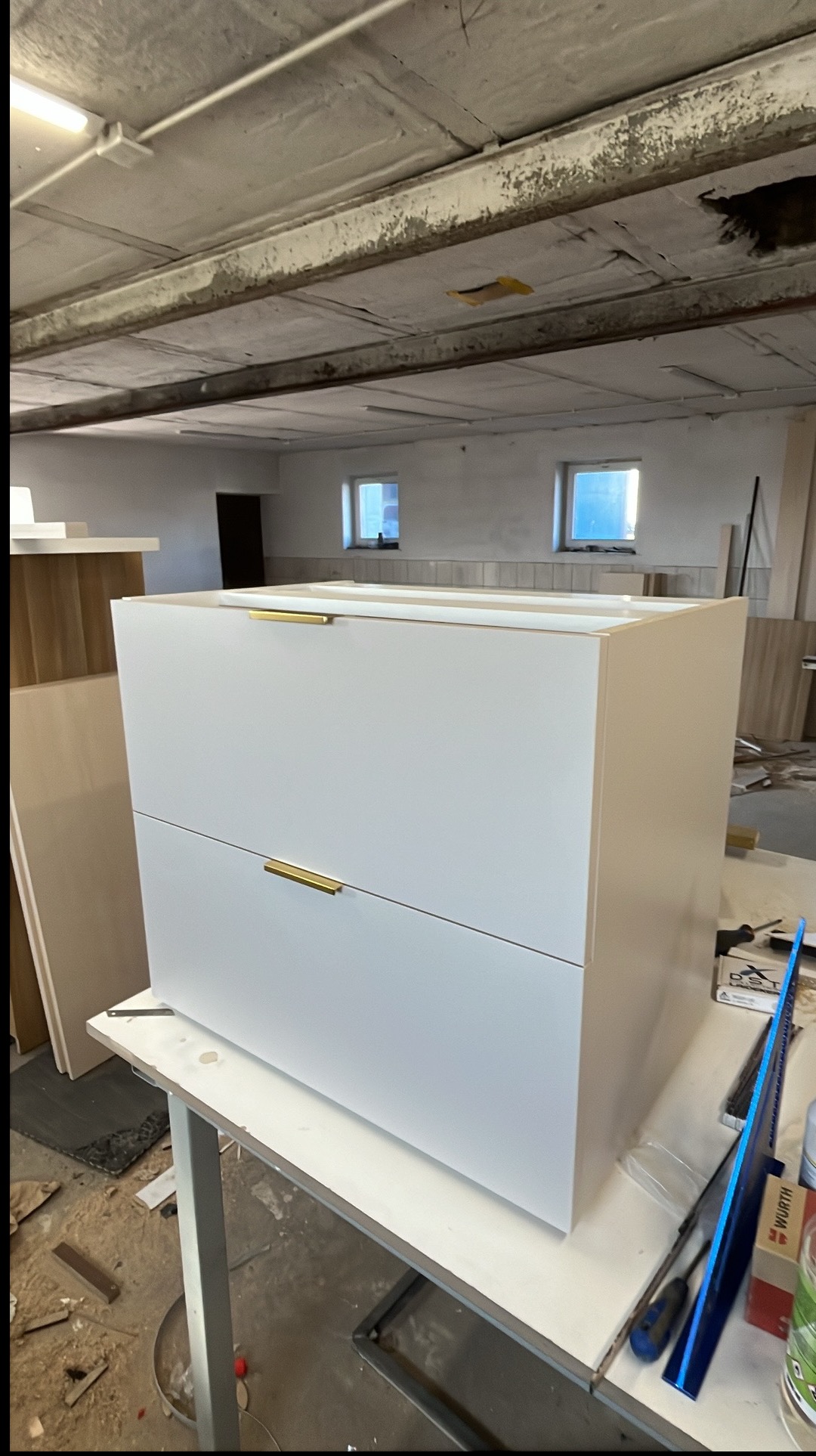 Biała szafka z dwoma szufladami i złotymi uchwytami, umieszczona na stole w warsztacie stolarskim, narzędzia widoczne w tle.