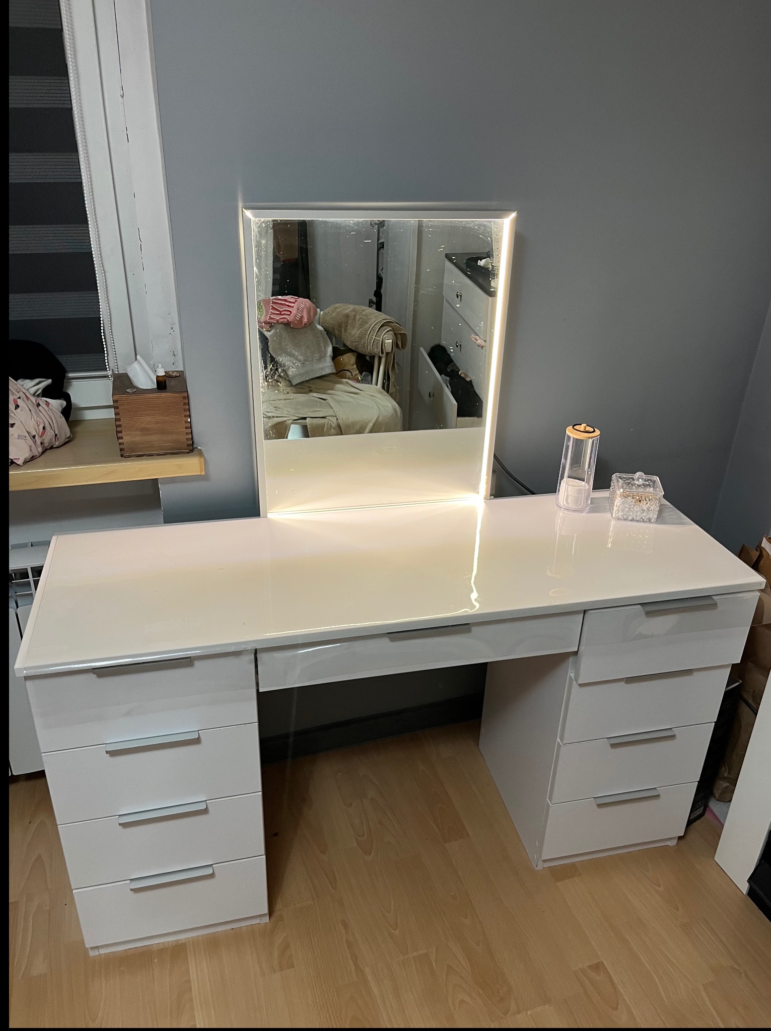 Biała toaletka z podświetlanym lustrem LED, ustawiona na drewnianej podłodze. Widoczne szuflady z metalowymi uchwytami i akcesoria na blacie.