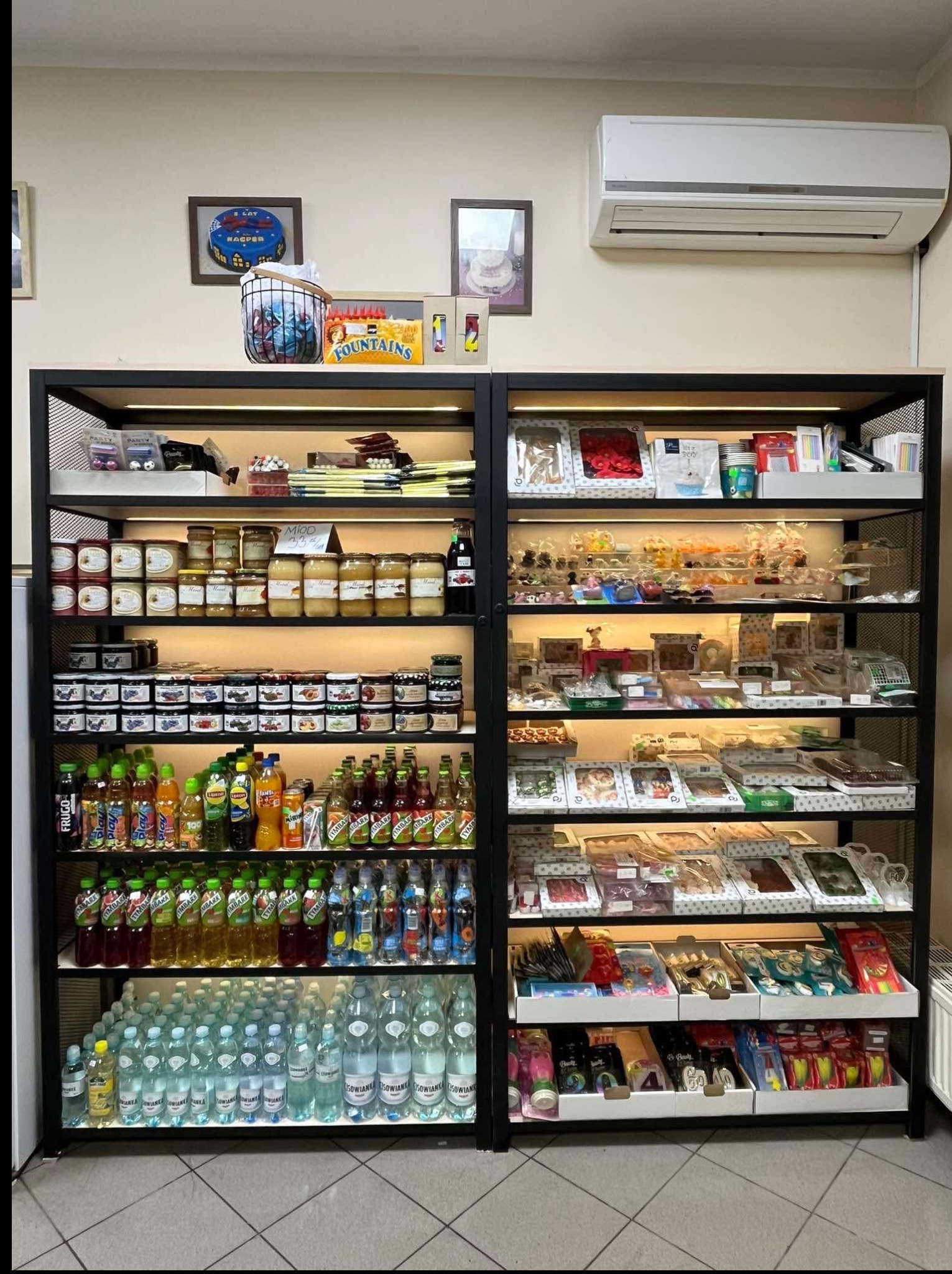 Regał sklepowy z metalową ramą, wypełniony słoikami z miodem, dżemami i napojami. Górne półki oświetlone, w tle klimatyzator i dekoracje ścienne.