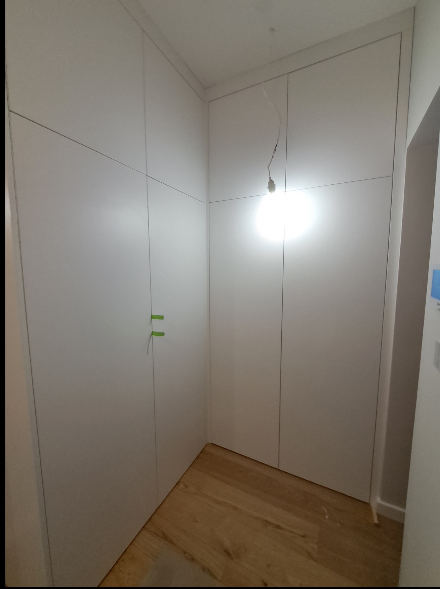 Wnętrze z zabudową szafy wnękowej z białymi frontami, widoczne zielone taśmy zabezpieczające. Na podłodze panele drewniane. Oświetlenie punktowe.