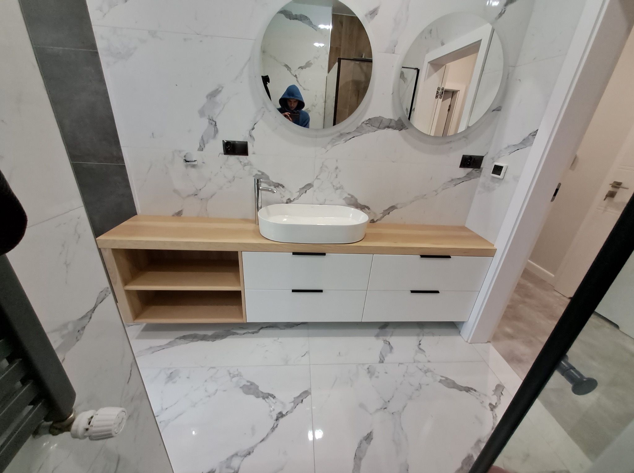 Nowoczesna łazienka z drewnianym blatem, białą umywalką i szafkami z czarnymi uchwytami. Dwa okrągłe lustra na ścianie z marmurowym wzorem.