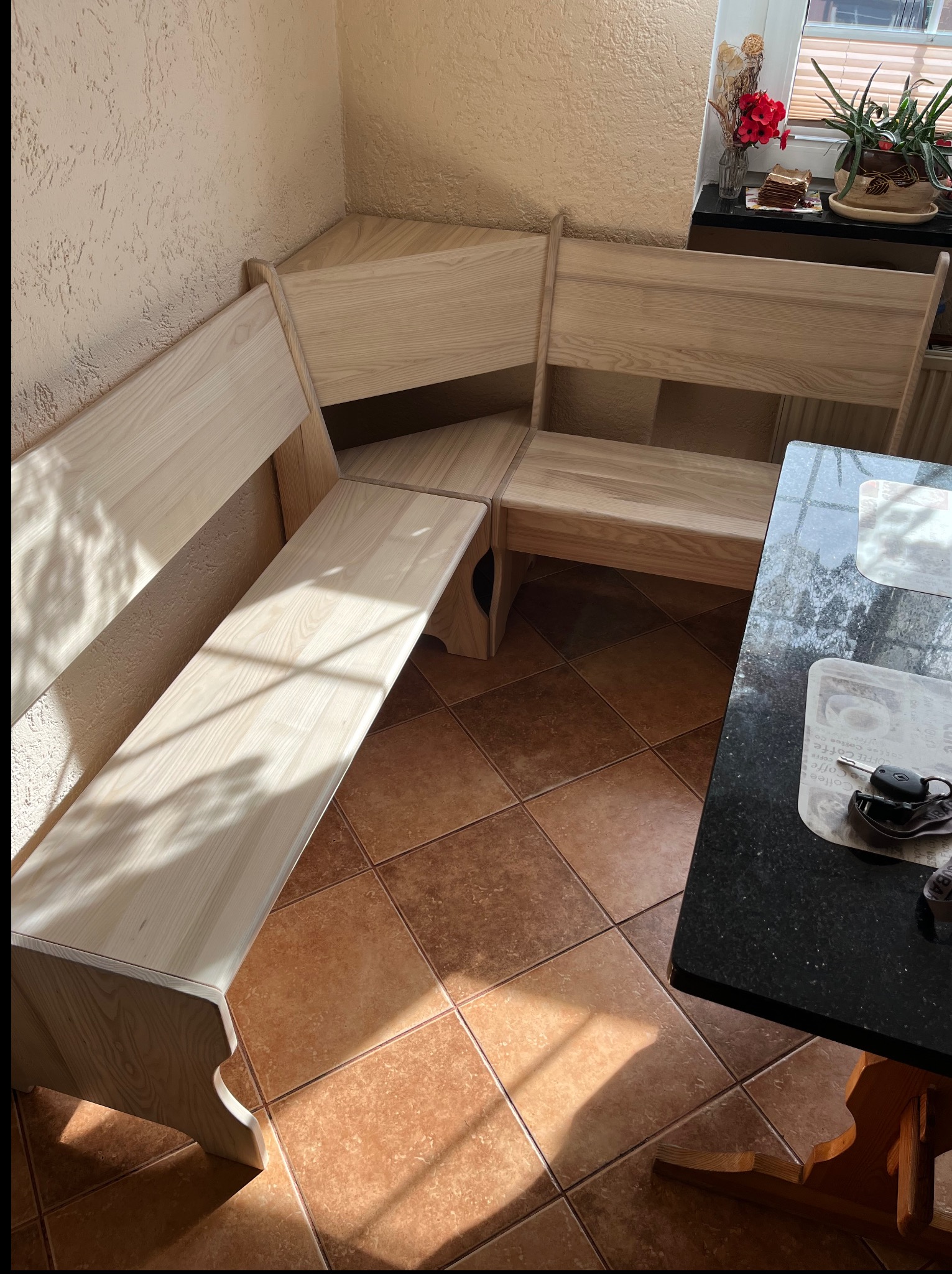 Jasna, drewniana ława narożna z oparciem, ustawiona w rogu pomieszczenia z terakotą. Obok stoi stół z czarnym blatem i kluczyki samochodowe.