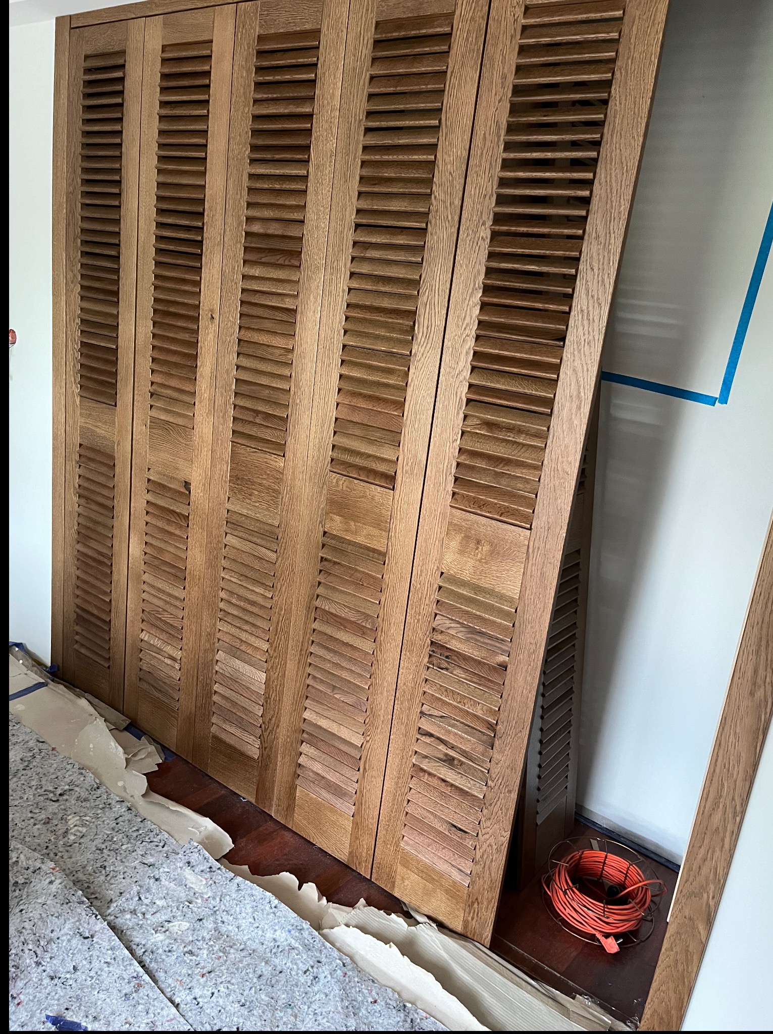 Drewniane, brązowe, lamelowe drzwi szafy wnękowej w trakcie montażu, oparte o ścianę z widocznymi narzędziami i zabezpieczeniami podłogi. Detal montażu.