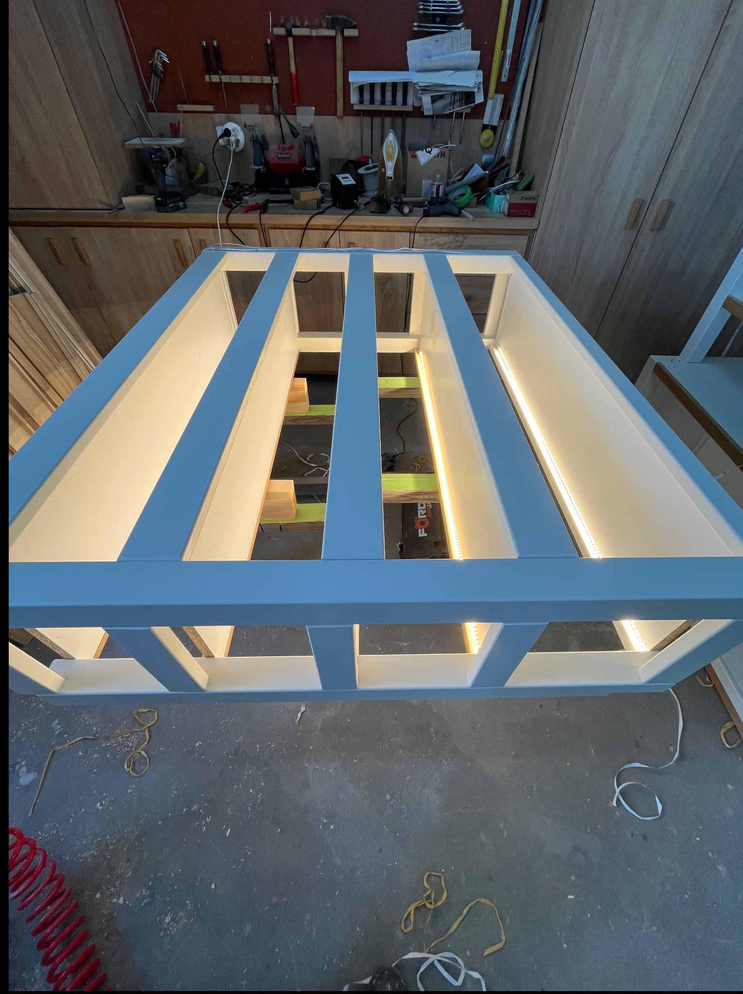 Biała konstrukcja meblowa w warsztacie, z wbudowanym oświetleniem LED, widok od dołu, narzędzia i akcesoria stolarskie w tle, surowy beton na podłodze.