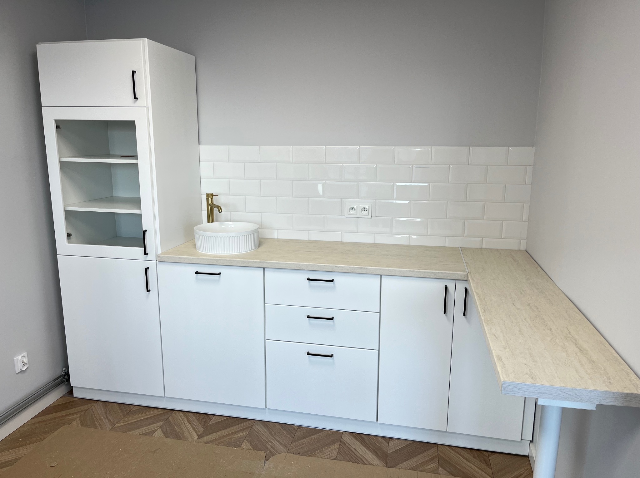 Biała zabudowa kuchenna z szafkami, blatem imitującym marmur i okrągłą umywalką. Minimalistyczne uchwyty, białe płytki na ścianie i drewniana podłoga w jodełkę.