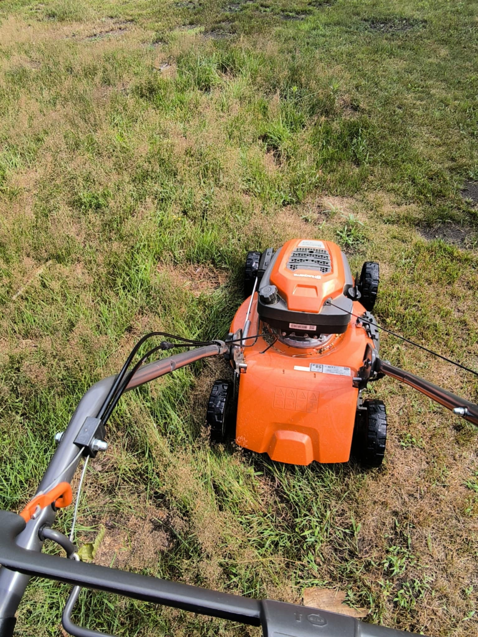 Pomarańczowa kosiarka Husqvarna na trawniku z perspektywy operatora. Widok z góry na urządzenie i trawę, z uchwytem w dolnej części kadru.