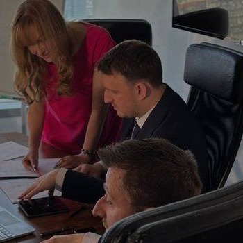 Zespół w biurze: kobieta sprawdza dokumenty, mężczyźni siedzą przy biurku, jeden z tabletem. Oświetlenie naturalne, jasne kolory. Praca zespołowa, analiza dokumentów.