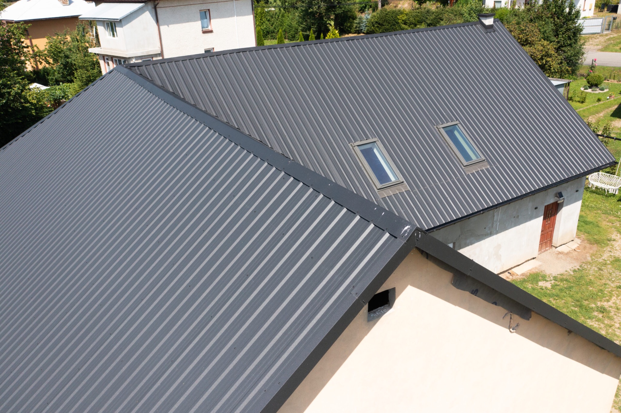 Szary dach z blachy trapezowej z dwoma oknami dachowymi, widoczny z góry. Budynek gospodarczy z czerwoną stolarką w tle. Dom w otoczeniu zieleni.