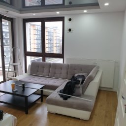 Adomex - Jasne wnętrze po remoncie: sofa z listwami, odnowiona podłoga, sufit z połyskiem i okna z widokiem na miasto. Widoczne ślady prac wykończeniowych.