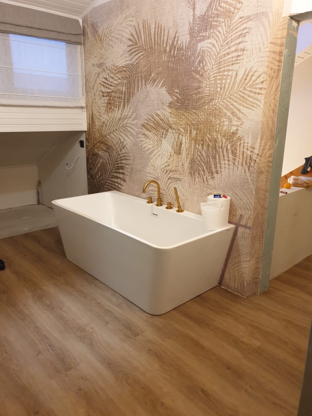 Biała, prostokątna wanna z złotą armaturą na tle ściany w palmy w łazience z panelami podłogowymi. Widoczne materiały wykończeniowe.