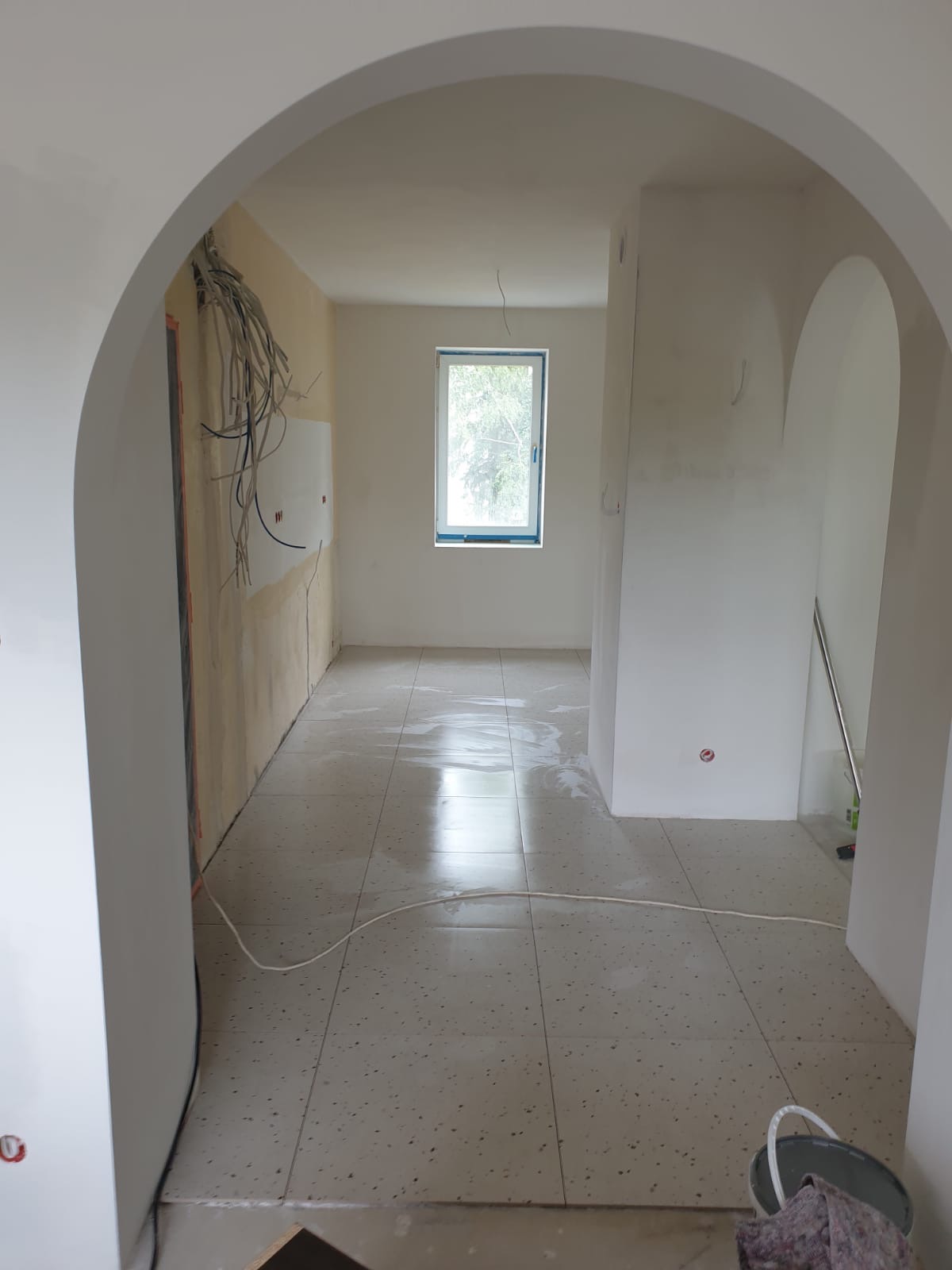 Wnętrze w trakcie remontu z łukowatymi przejściami, świeżo położone płytki podłogowe, widoczne kable i przygotowania do malowania ścian. Okno z widokiem na zieleń.
