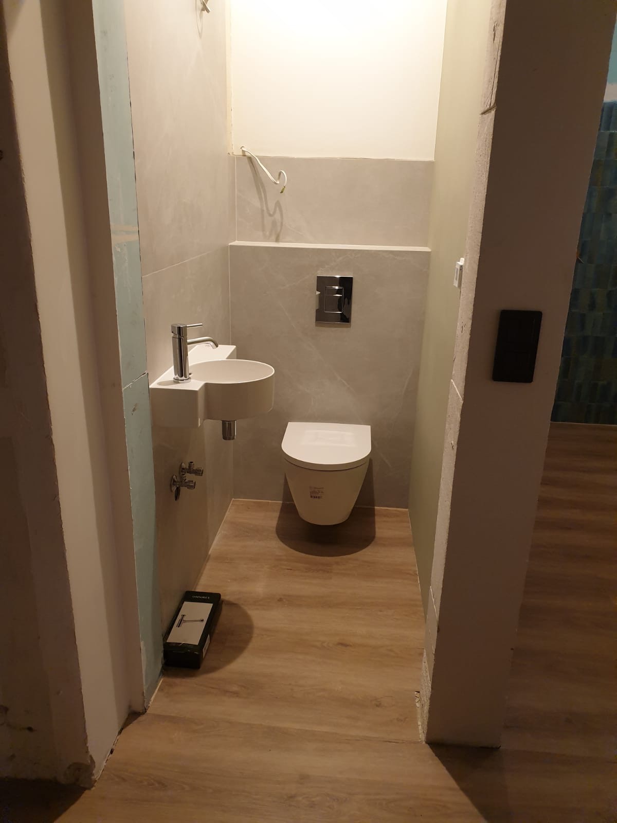 Nowoczesna, wykończona łazienka z podwieszaną toaletą, umywalką na blacie i drewnopodobną podłogą. Widoczne elementy instalacji i pudełko narzędzi.