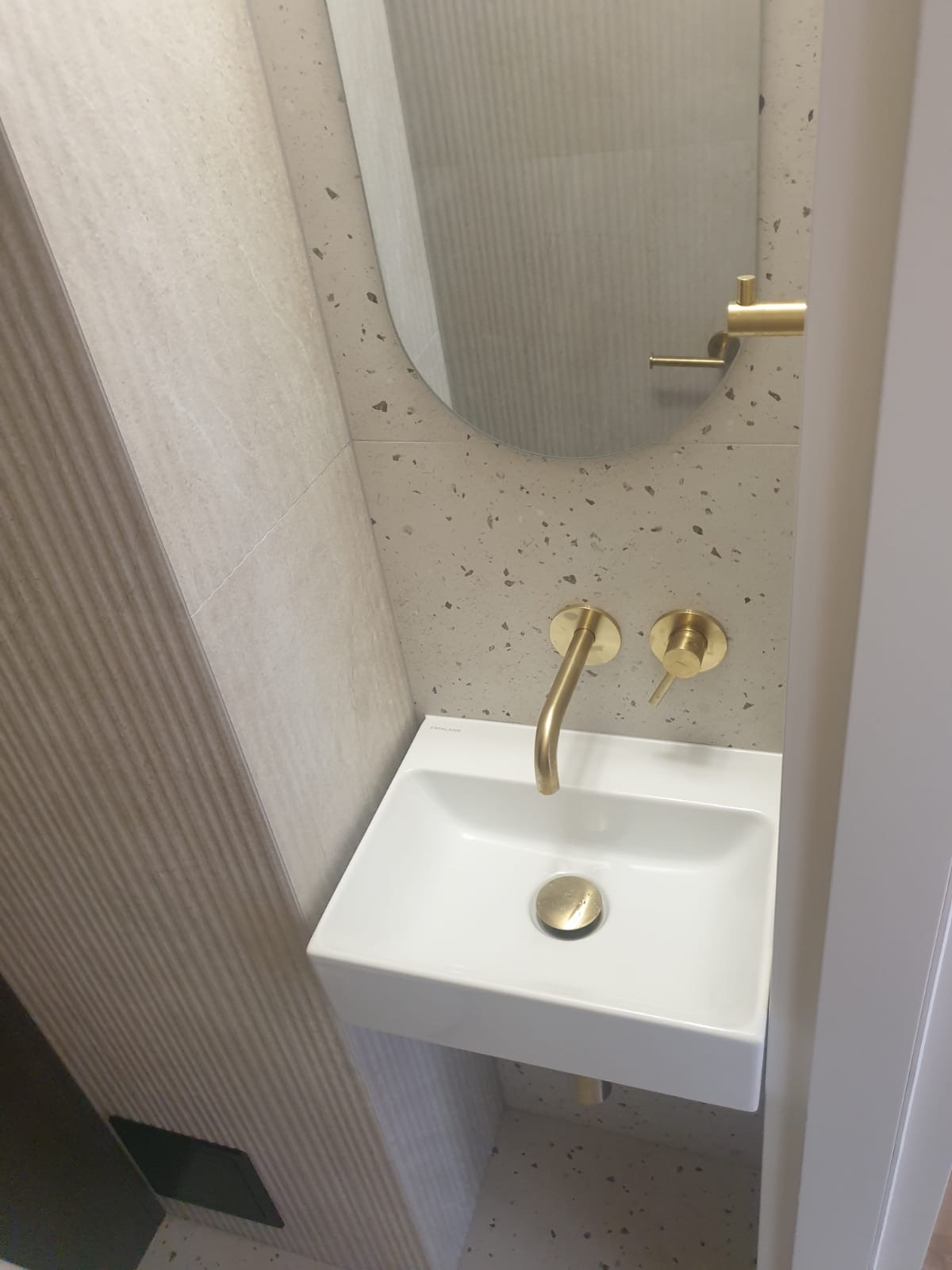 Nowoczesna, minimalistyczna łazienka w Warszawie: biała umywalka z złotą armaturą na tle ściany z drobnymi plamkami i owalne lustro.