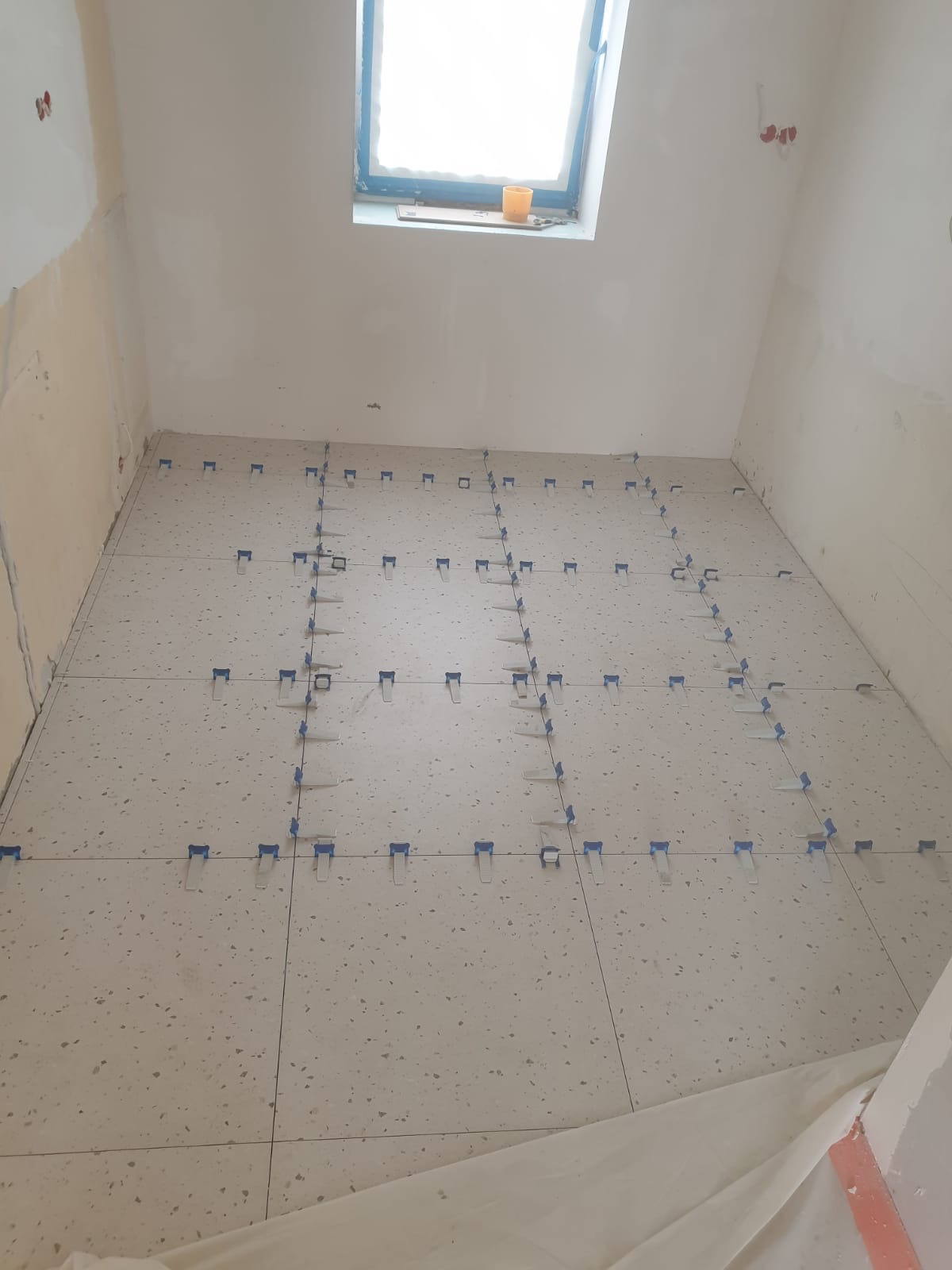 Układanie płytek podłogowych w łazience na poddaszu, z użyciem klipsów poziomujących. Widoczne okno dachowe i surowe ściany w trakcie remontu.