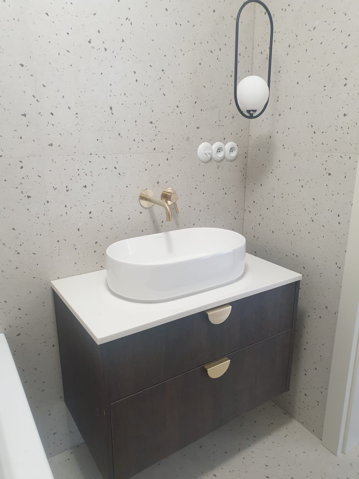 Nowoczesna łazienka: biała umywalka nablatowa na szafce z ciemnego drewna, złota bateria, ściana i podłoga w płytkach lastryko, minimalistyczna lampa.