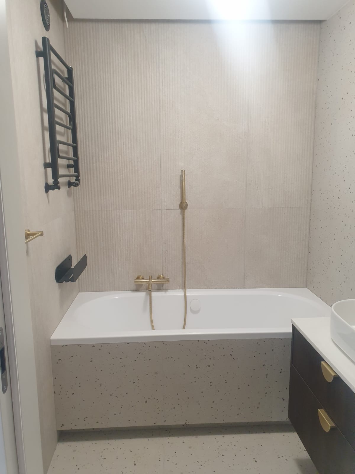 Nowoczesna łazienka z wanną obudowaną płytkami lastryko, złota armatura, grzejnik drabinkowy i umywalka z szafką. Ściany wyłożone płytkami w beżowych odcieniach.