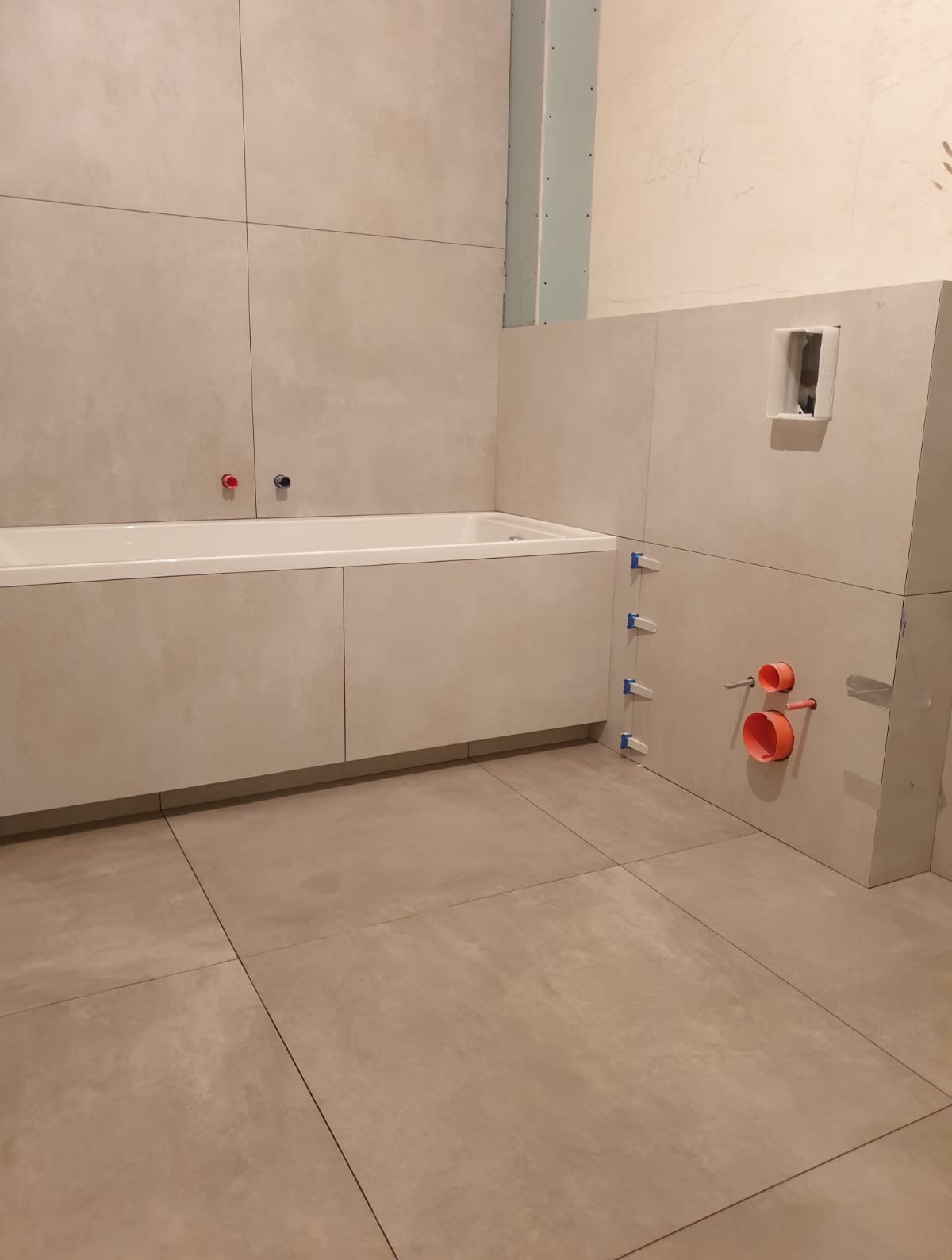 Łazienka w trakcie wykańczania: beżowe płytki na ścianach i podłodze, zabudowana wanna, instalacja wodna z pomarańczowymi rurami. Widoczne krzyżyki glazurnicze.