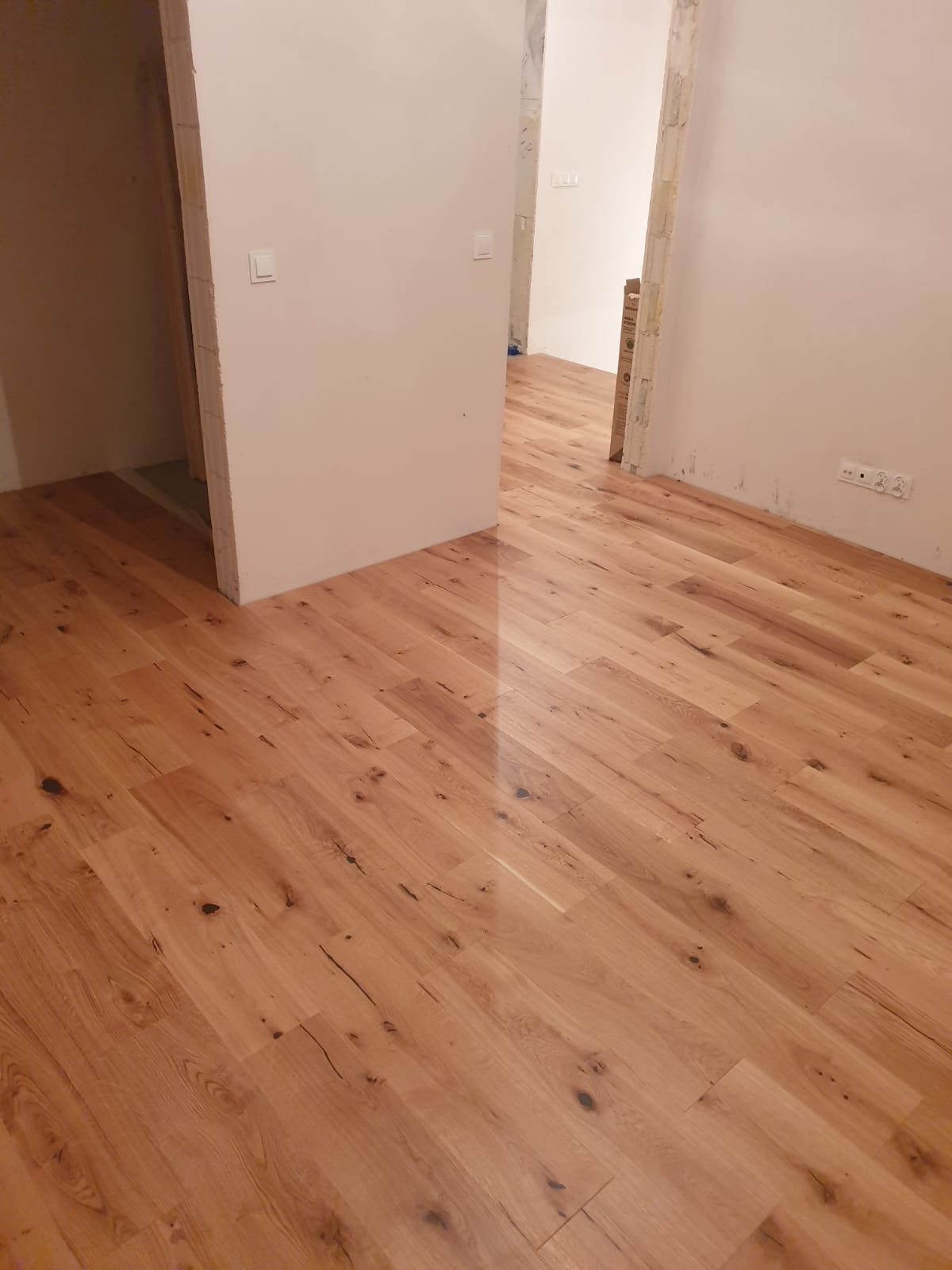 Ukończona podłoga z paneli drewnianych w jasnym odcieniu, widoczna w surowym wnętrzu z białymi ścianami i przygotowanymi otworami drzwiowymi.