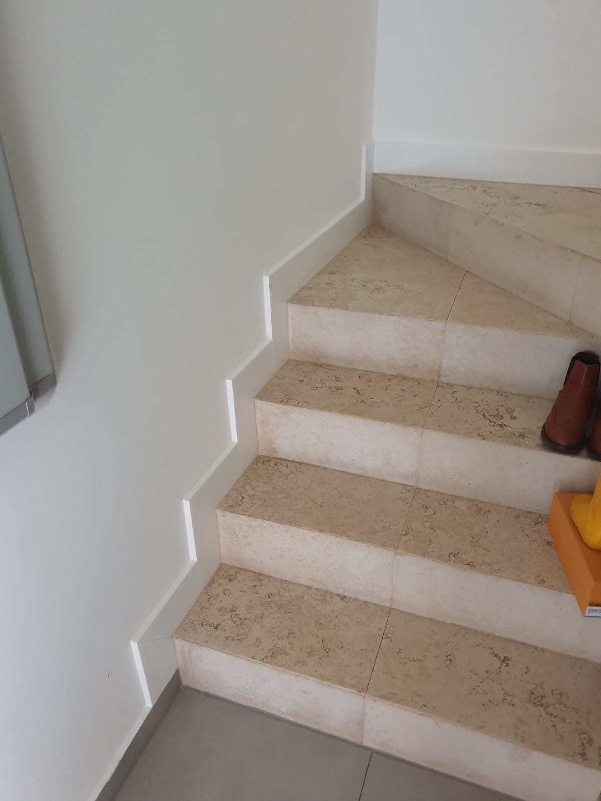 Wykończone schody z beżowych płytek, białe listwy przypodłogowe. U góry widoczne buty i pudełko. Detal wykończenia wnętrza.