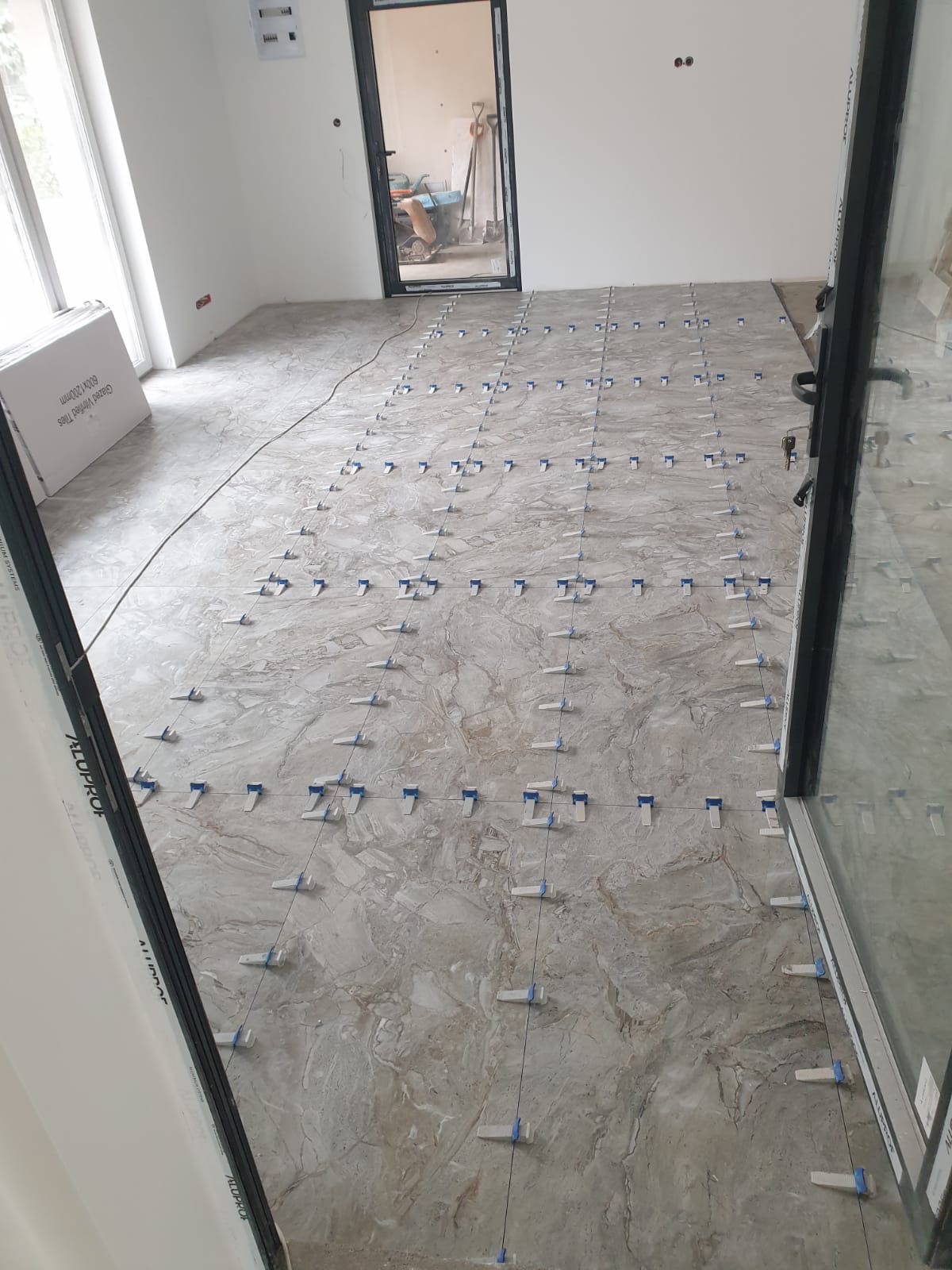 Układanie płytek podłogowych w jasnym wnętrzu. Widoczne krzyżyki dystansowe i paczka płytek przy ścianie. Praca w toku, jasne płytki imitujące marmur.