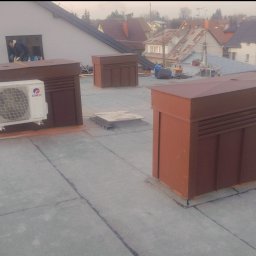 FlachDach - Płaski dach z brązowymi obudowami wentylacyjnymi i klimatyzatorem Gree. W tle zabudowa jednorodzinna i osoba pracująca w oknie budynku.