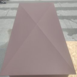 FlachDach - Brązowy komin wentylacyjny na dachu z szarych płyt, widok z góry. W tle dom i drzewa. Komin ma geometryczny kształt i jest głównym elementem zdjęcia.