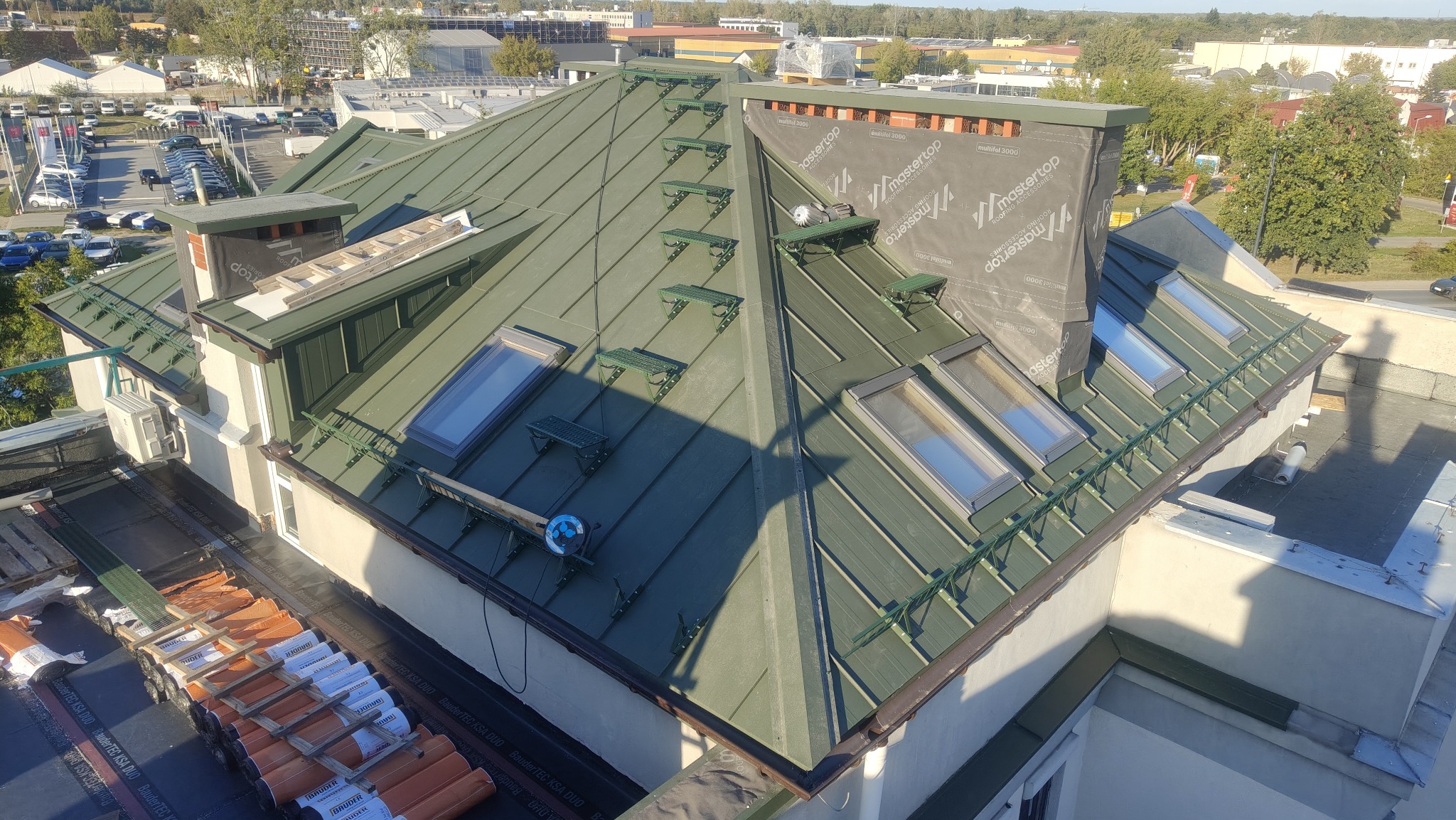Zielony dach z blachy trapezowej z oknami dachowymi i stopniami kominiarskimi. Częściowo zakryty komin. Na dole widoczne materiały budowlane. Ujęcie z góry.