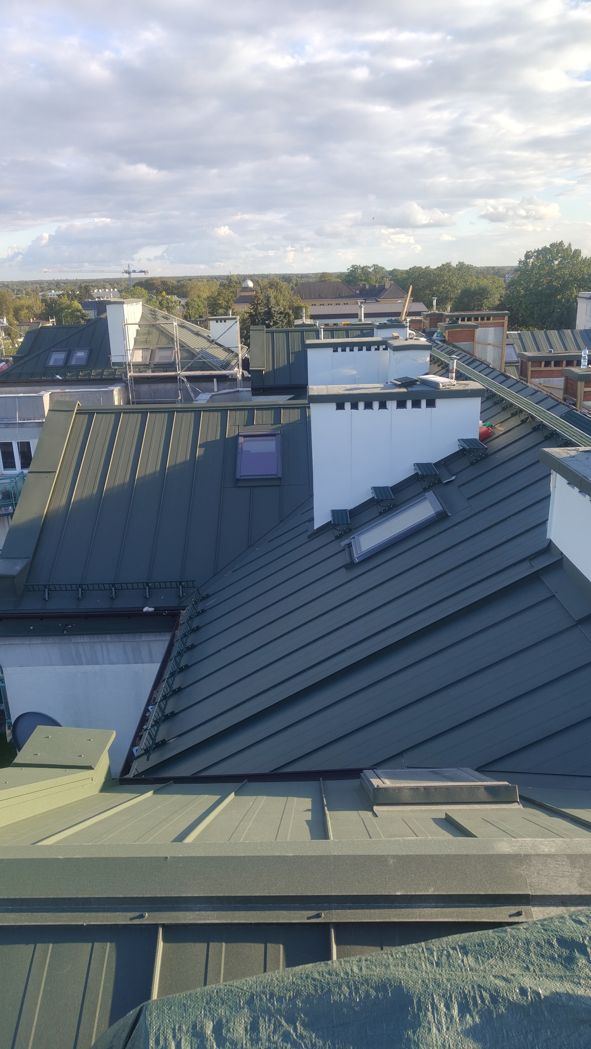 Szary dach z blachy trapezowej, z oknami dachowymi i kominami wentylacyjnymi. Widok z góry na dachy budynków w oddali i błękitne niebo z chmurami.