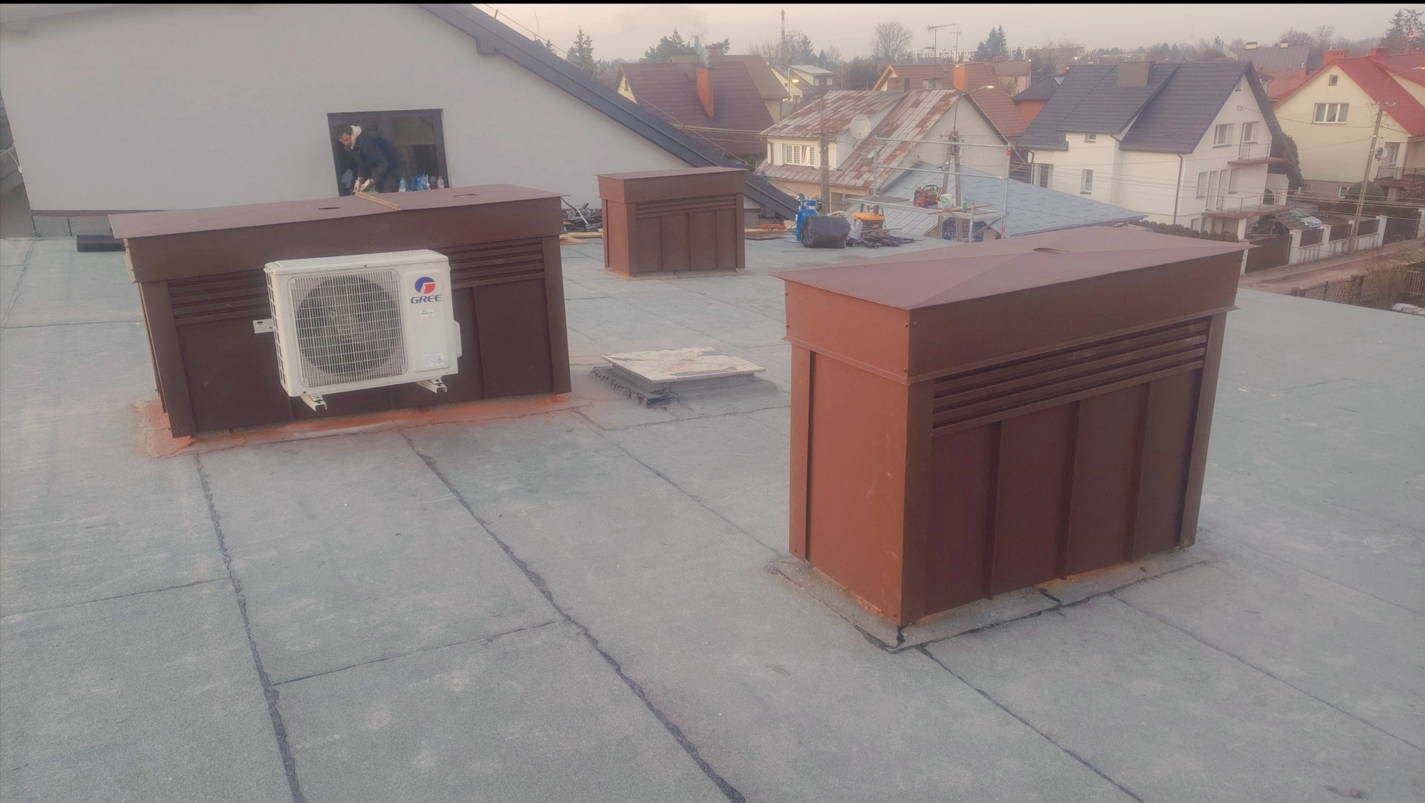 Płaski dach z brązowymi obudowami wentylacyjnymi i klimatyzatorem Gree. W tle zabudowa jednorodzinna i osoba pracująca w oknie budynku.