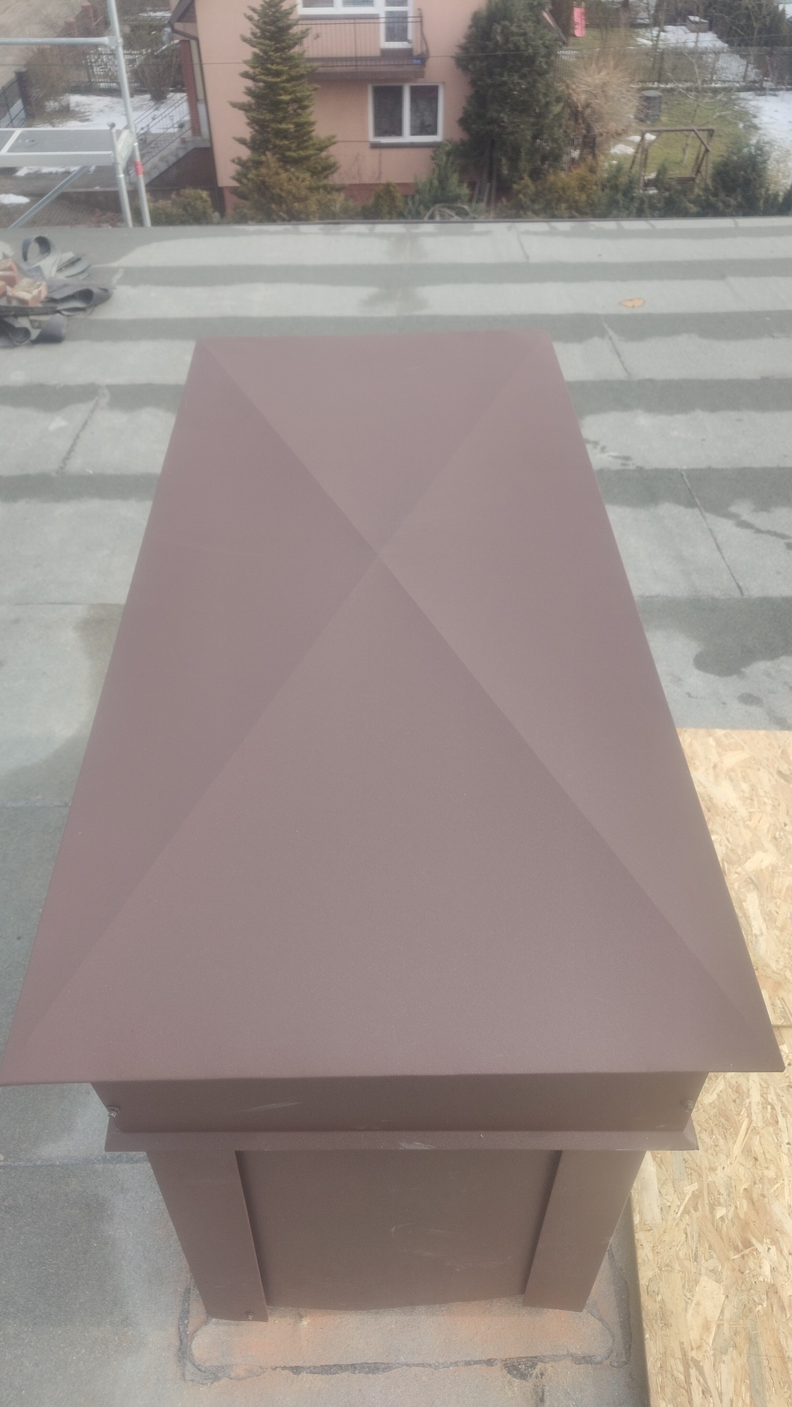 Brązowy komin wentylacyjny na dachu z szarych płyt, widok z góry. W tle dom i drzewa. Komin ma geometryczny kształt i jest głównym elementem zdjęcia.