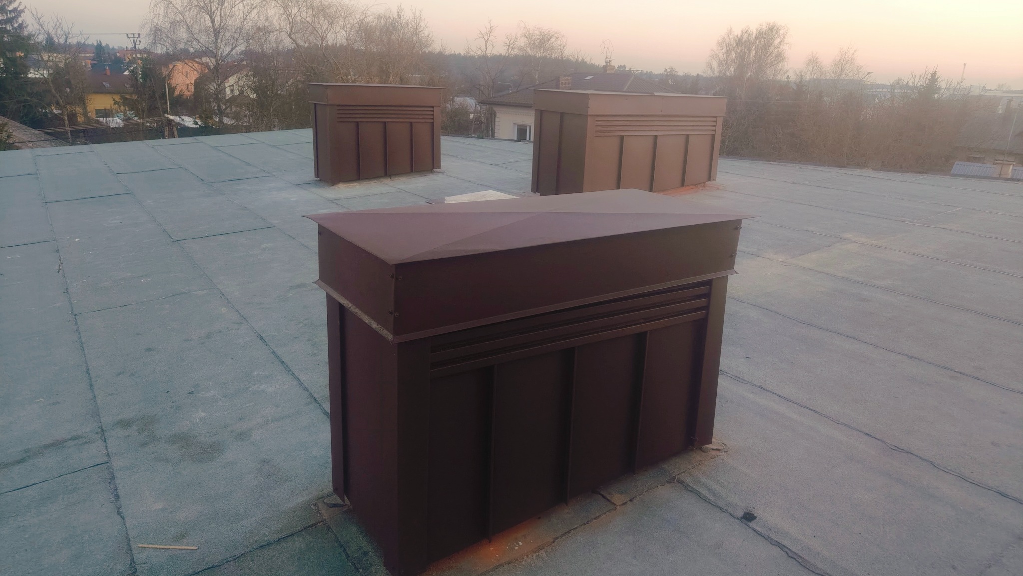 Płaski dach pokryty papą z trzema brązowymi kominami wentylacyjnymi. Widok z góry, w tle zabudowa jednorodzinna i drzewa.