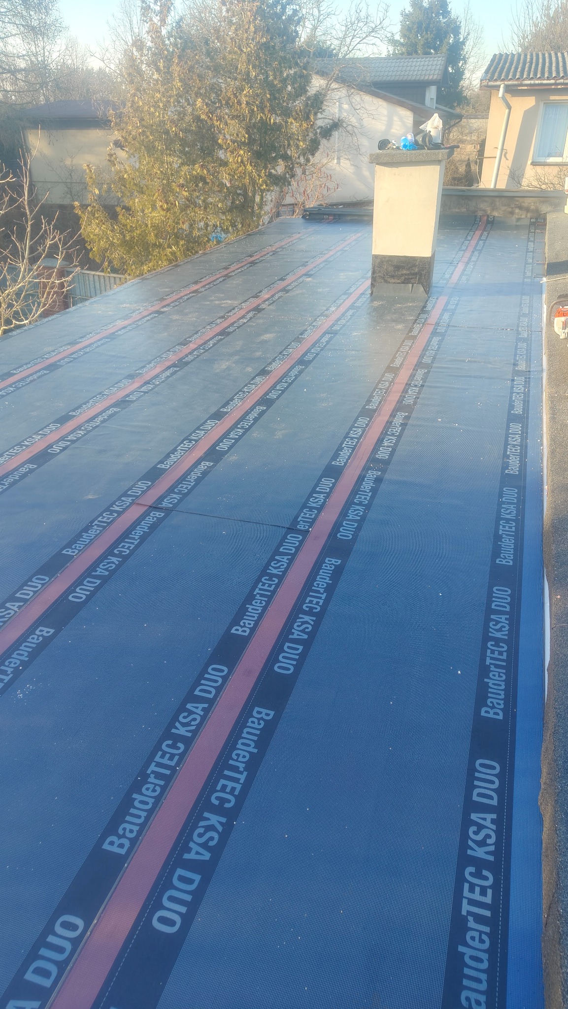 Dach płaski pokryty papą termozgrzewalną BauderTEC KSA DUO, z widocznym kominem i elementami otoczenia. Układanie papy w pasach z zachowaniem zakładek.