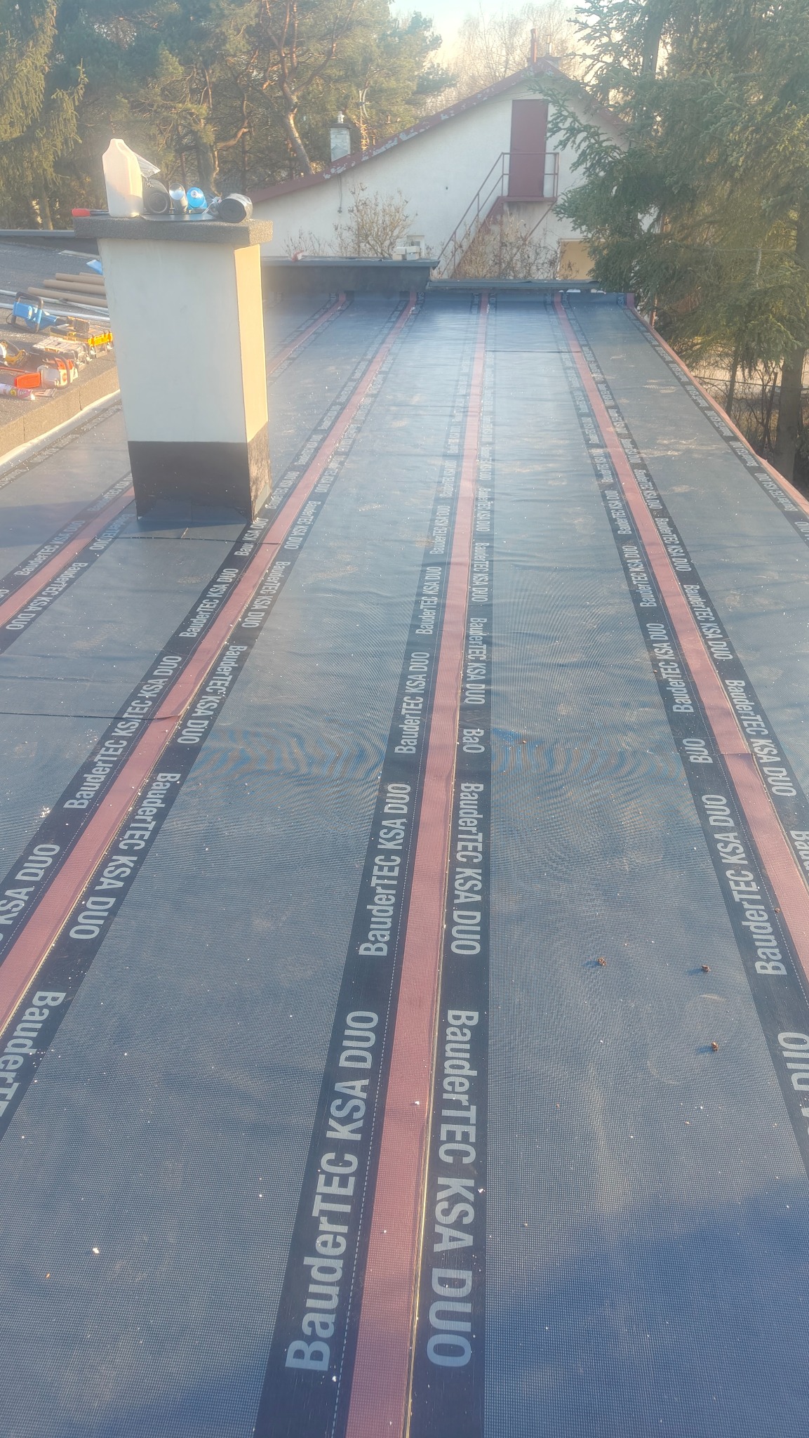 Dach płaski pokryty papą z widocznymi pasami z nazwą producenta, komin na dachu, w tle budynek i drzewa w słoneczny dzień. Detal hydroizolacji.