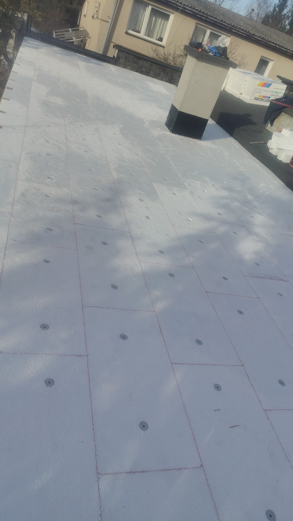 Ocieplenie dachu styropianem z widocznymi łącznikami mechanicznymi. Fragment komina i budynku w tle. Prace dekarskie w trakcie realizacji.