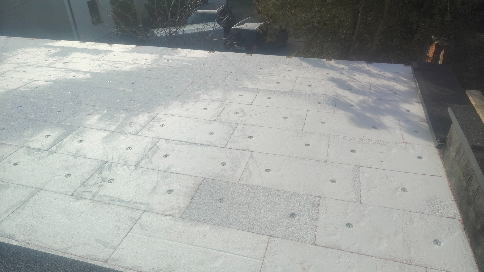 Białe płyty izolacyjne, prawdopodobnie styropian, ułożone na dachu i przymocowane metalowymi kołkami. Widok z góry, fragment dachu i otoczenia.