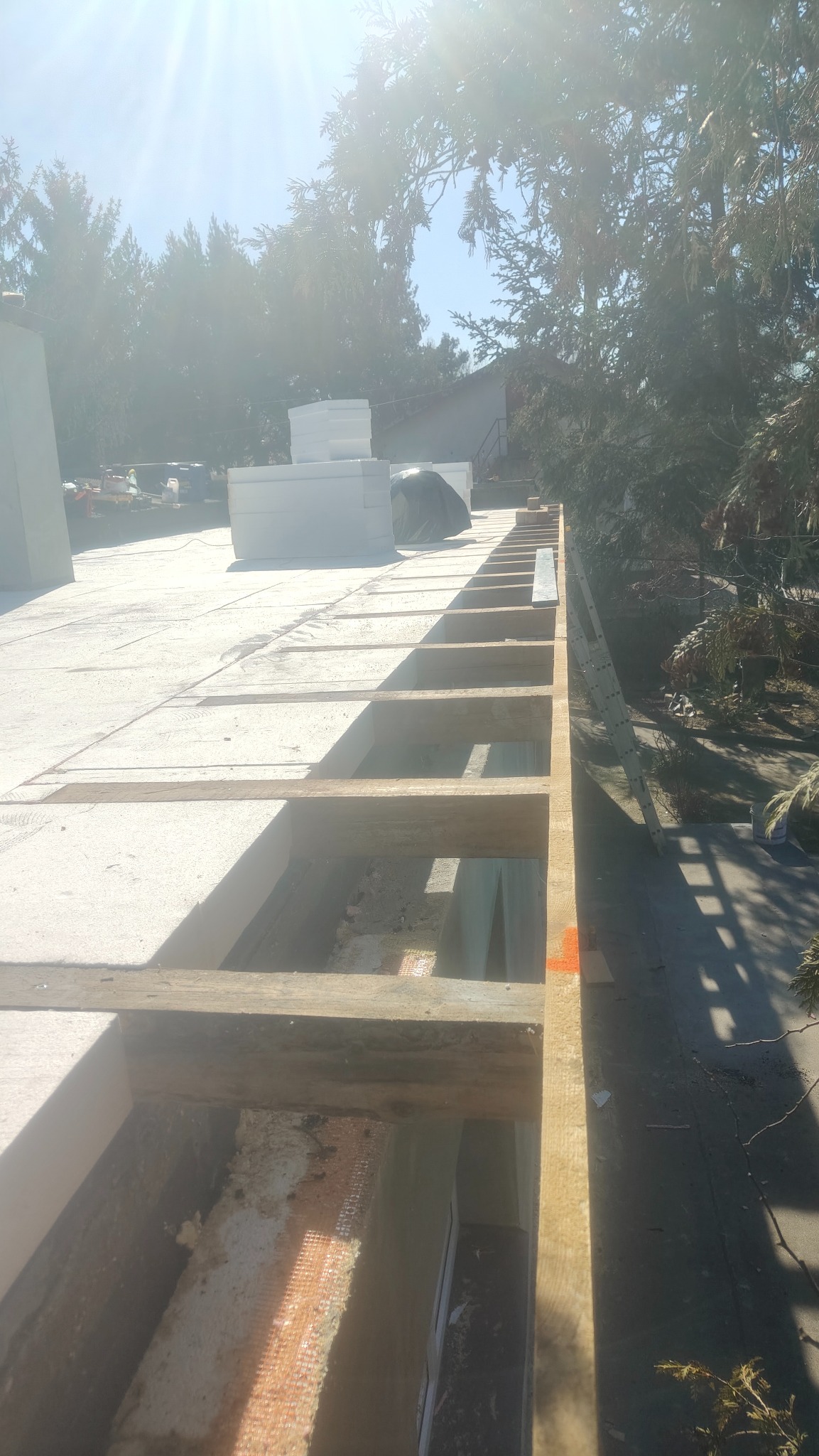 Wysoka perspektywa na konstrukcję dachu z drewnianymi belkami i izolacją, w słoneczny dzień. Widoczne elementy więźby i ocieplenia, drzewa w tle.