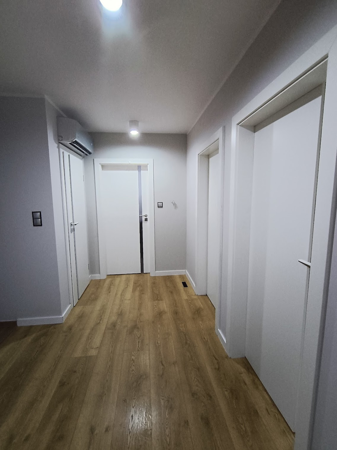 Nowoczesny, minimalistyczny przedpokój z klimatyzatorem na ścianie. Parkiet w odcieniach brązu, białe ściany i drzwi. Widoczny fragment otwartych drzwi do pokoju.