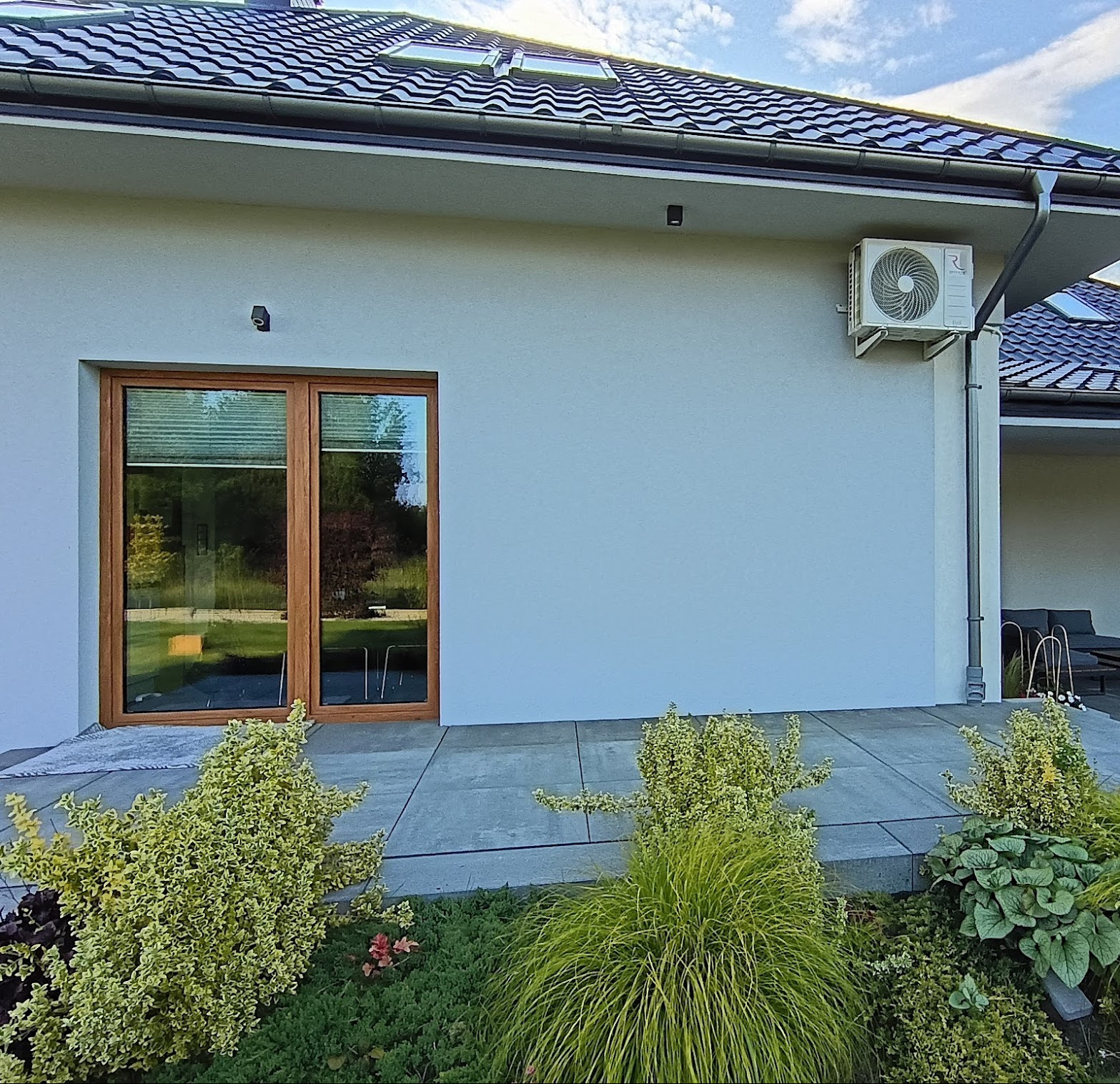 Biała elewacja domu z zamontowaną klimatyzacją zewnętrzną, widok z poziomu ogrodu. Okno z drewnianą ramą. Dach z dachówką.