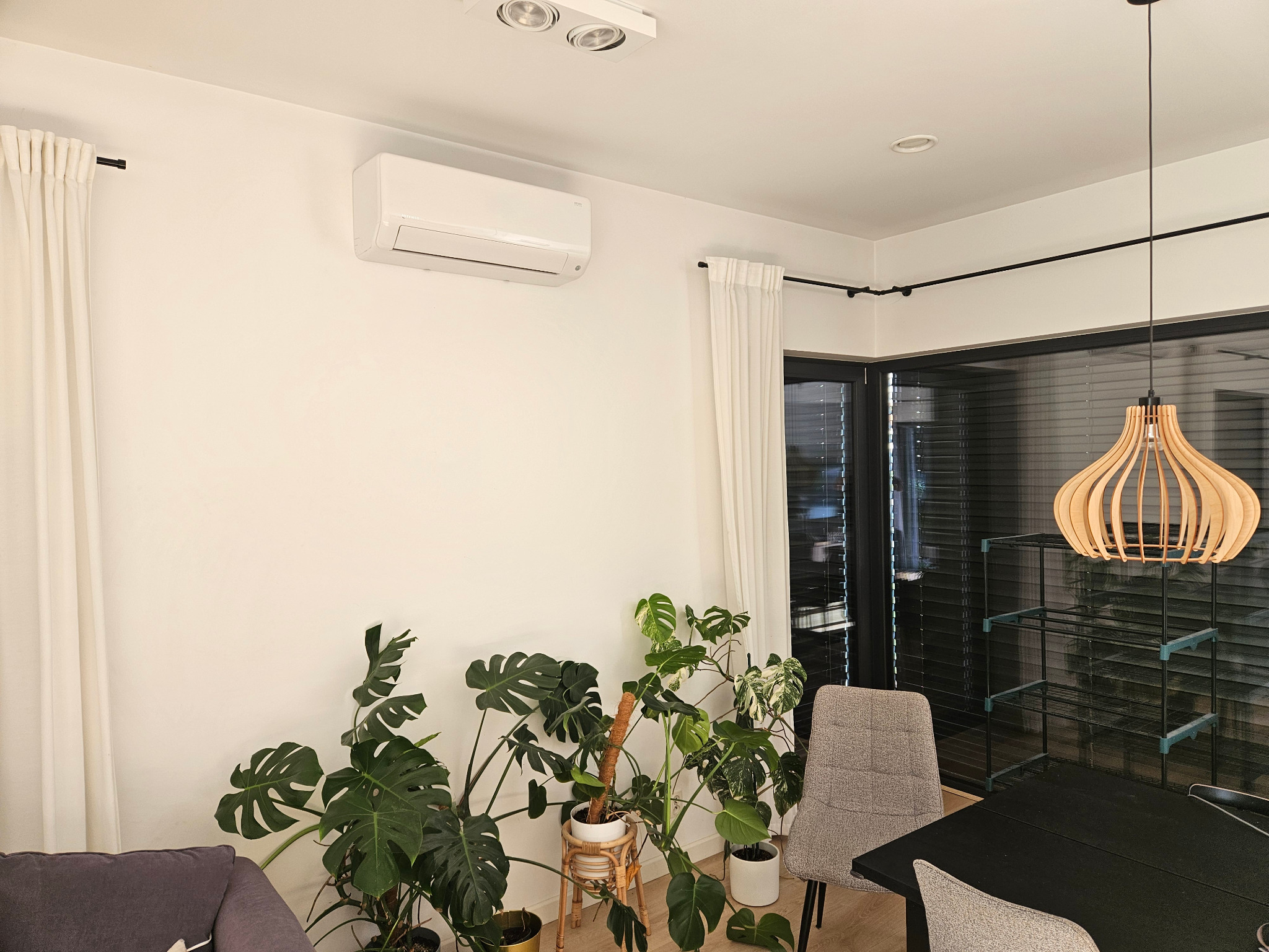 Nowoczesne wnętrze z klimatyzatorem ściennym, białymi zasłonami i roślinami doniczkowymi. Minimalistyczny design, jasne ściany i drewniana lampa wisząca.