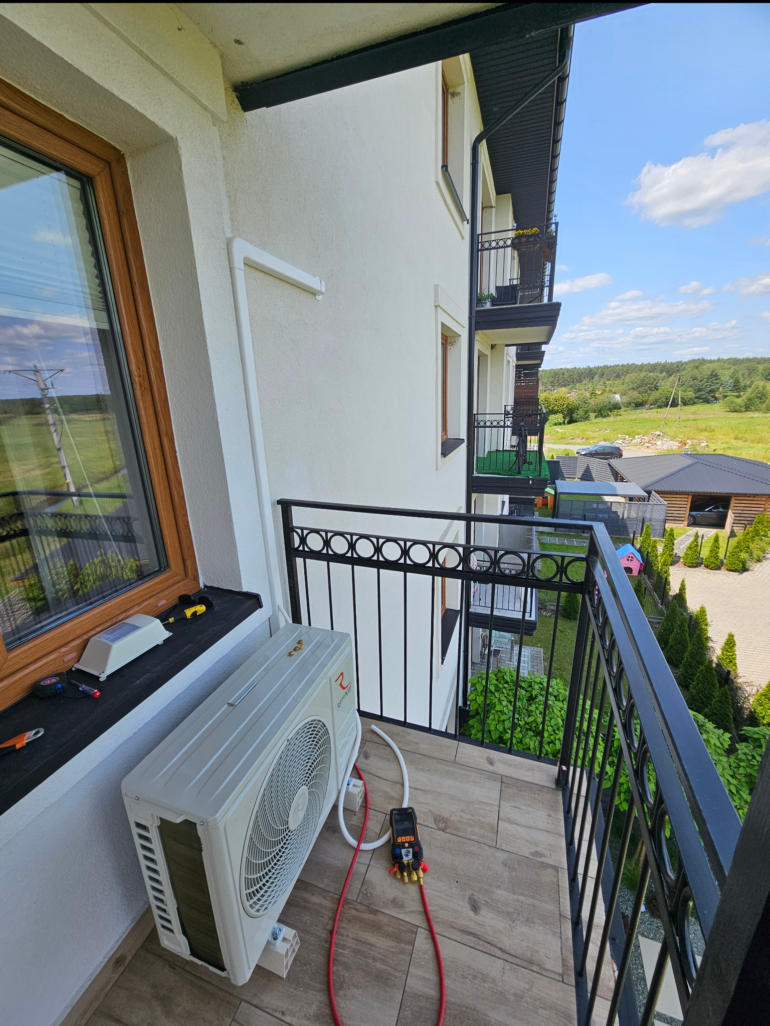 Biała jednostka zewnętrzna klimatyzacji na balkonie z czarną balustradą. Widoczne narzędzia serwisowe i przewody. W tle budynek i krajobraz.