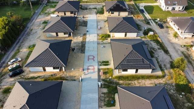 Widok z drona na osiedle nowych, parterowych domów z ciemnymi dachami, oddzielonych betonową drogą z czerwonym napisem. Częściowo zagospodarowany teren.