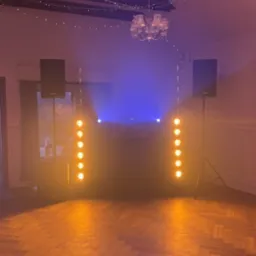 Scena z nagłośnieniem i oświetleniem w pomarańczowo-niebieskiej tonacji, dwa głośniki na statywach i kolumny świetlne po bokach, parkiet na podłodze, żyrandol na suficie.