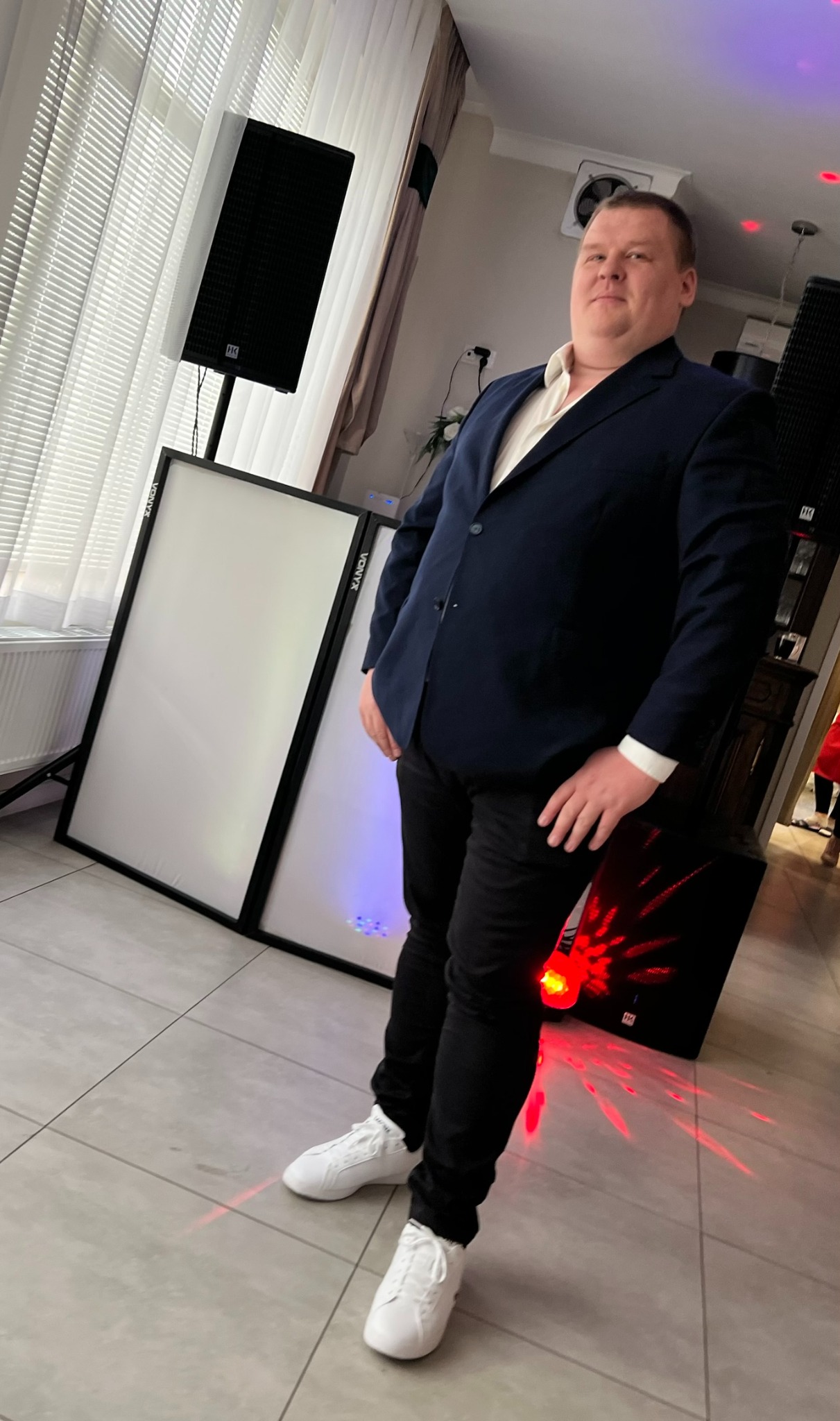 Mężczyzna w garniturze, białe panele oświetleniowe Vonyx, głośnik HK Audio i czerwone światła dyskotekowe tworzą scenę gotową na imprezę.