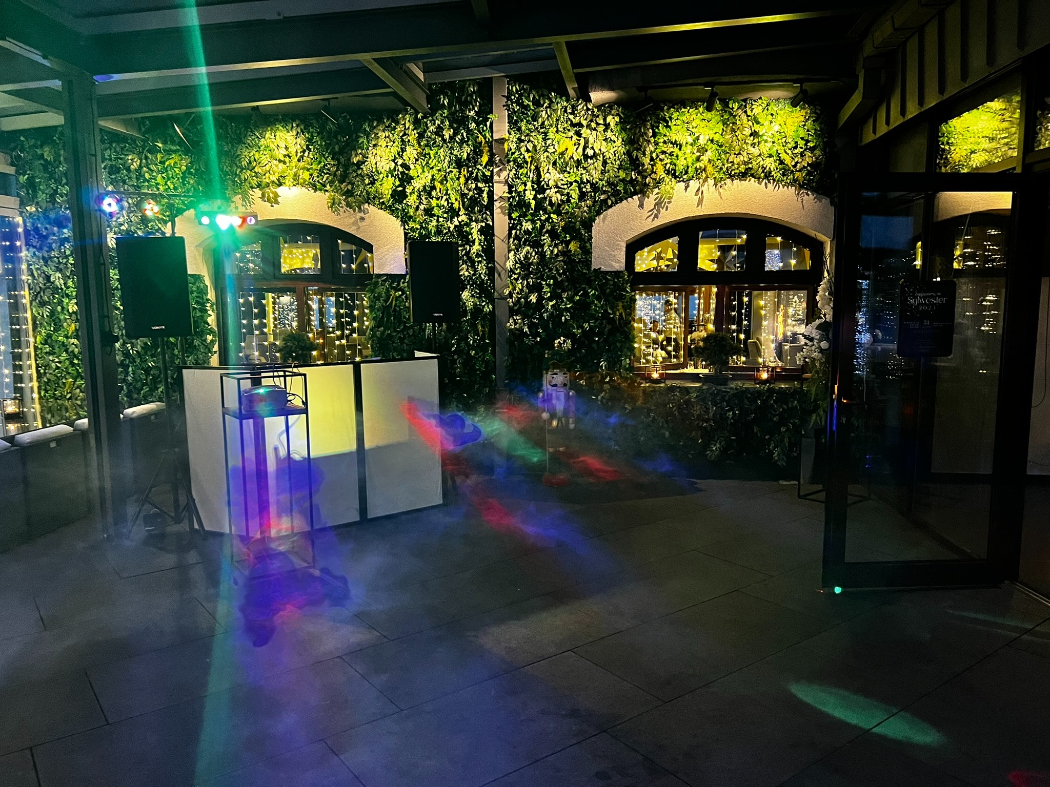 Sala z DJ-ką, nagłośnieniem i oświetleniem, dekoracją roślinną na ścianach oraz oknami z girlandami świetlnymi, przygotowana na imprezę sylwestrową.