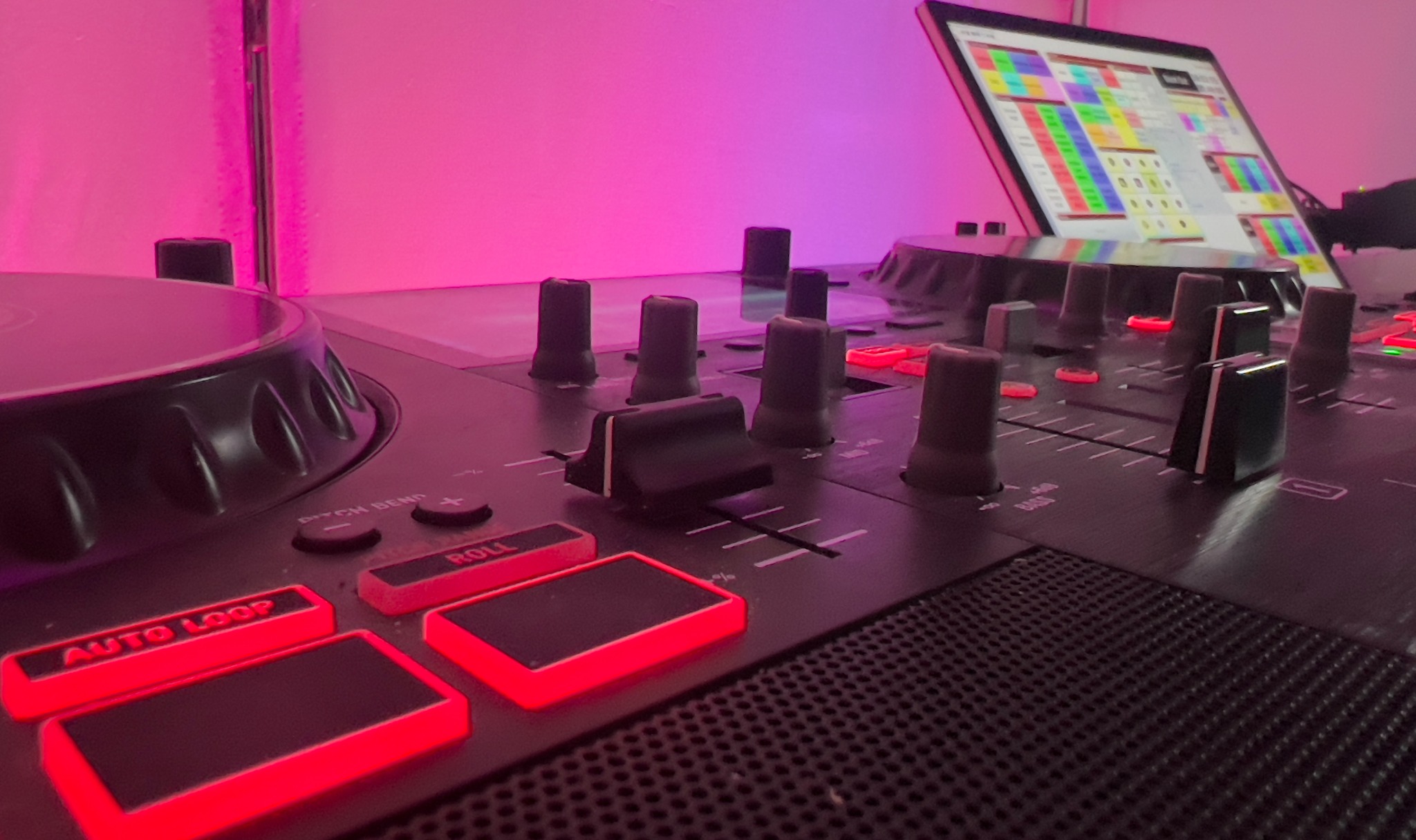 Czarny kontroler DJ z podświetlanymi na czerwono przyciskami i suwakami, ustawiony pod kątem, z widocznym ekranem laptopa w tle, oświetlony różowym światłem.
