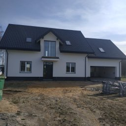 FASAD-MAX ROMAN ZDOROVYK - Biały dom jednorodzinny z ciemną dachówką i szarą podmurówką w słoneczny dzień. Widoczne rusztowania. Elewacja po renowacji.